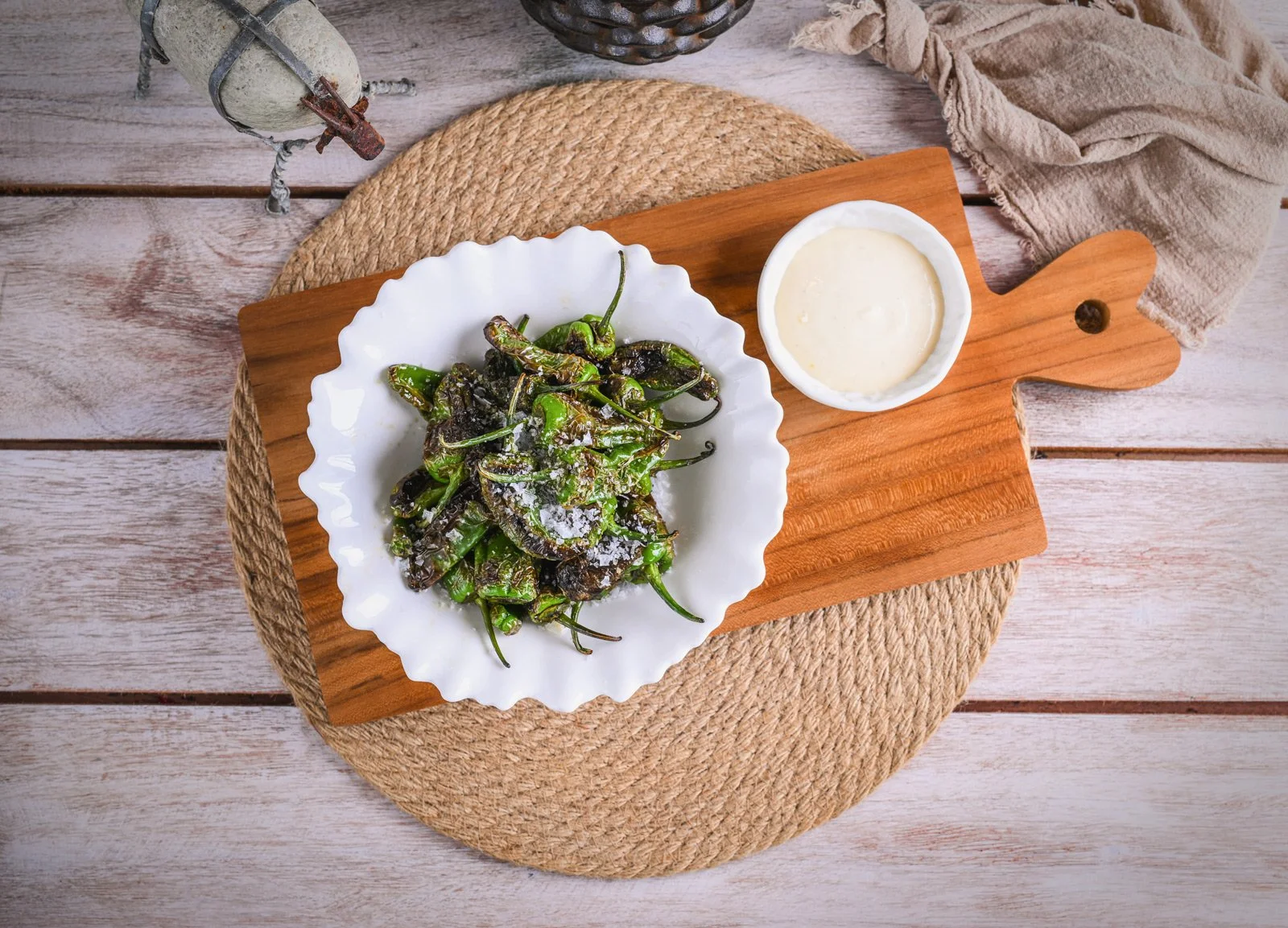 A white scalloped bowl filled with roasted green chilies sprinkled with salt, placed on a wooden serving board. Next to the bowl, there is a small dish of white dipping sauce. The setup is on a round woven placemat on a rustic wooden table with a bei