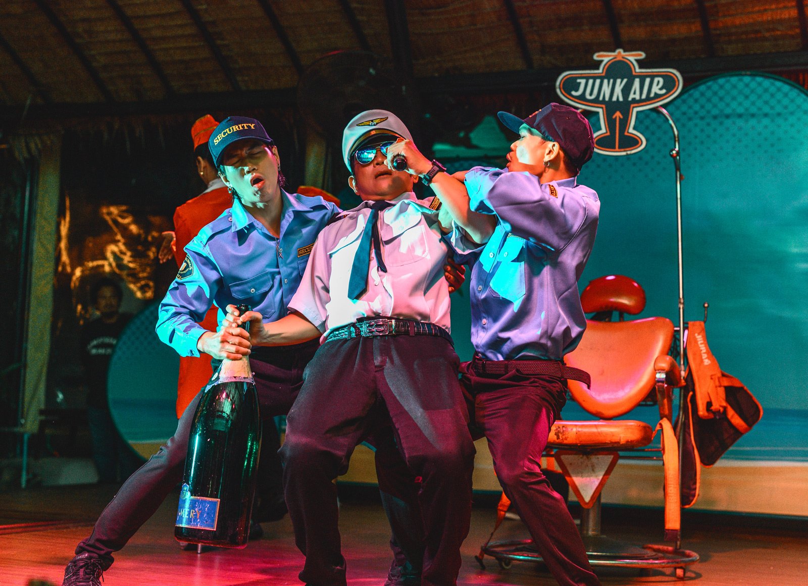 Three individuals dressed as security guards and a pilot on a stage, engaged in a lively performance. One person is holding a champagne bottle, another is being pushed with a microphone, and the third is assisting. There is a "Junk Air" sign and an o