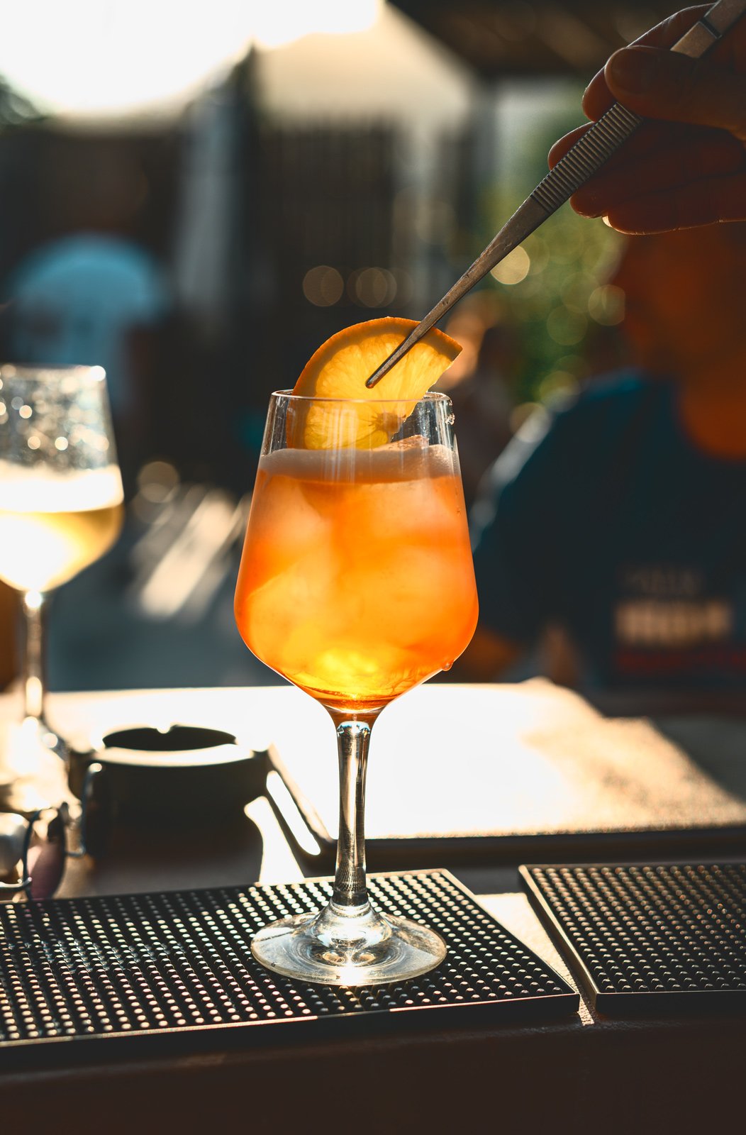 A cocktail glass filled with orange-colored drink and ice, garnished with an orange slice, sitting on a bar mat with sunlight in the background.