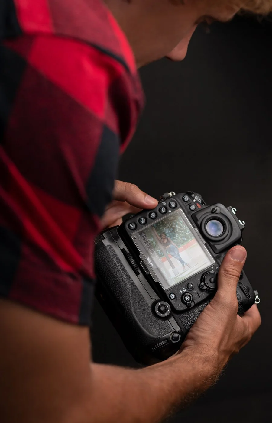 A person in a red and black plaid shirt holding a professional camera and looking at its display screen, which shows a photo of a woman posing outdoors.