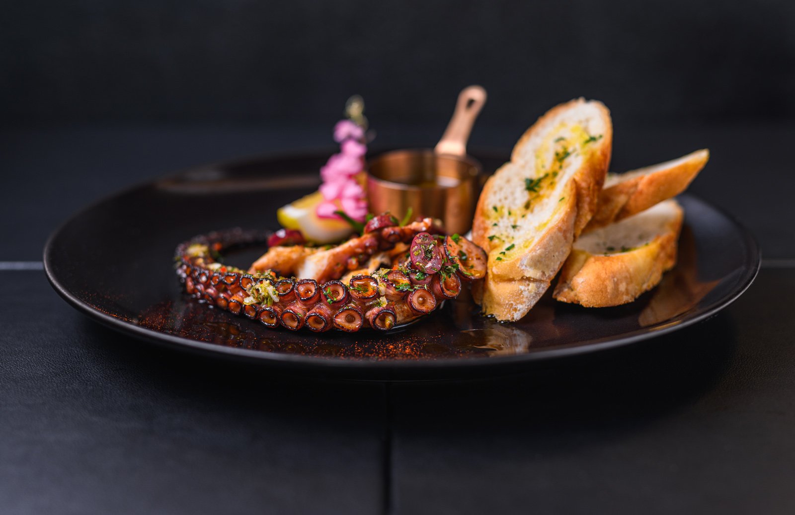 A black plate contains grilled octopus tentacle garnished with chopped herbs, accompanied by toasted bread slices with herb butter, and garnished with a small copper container of sauce, a lemon wedge, and pickled pink onions.