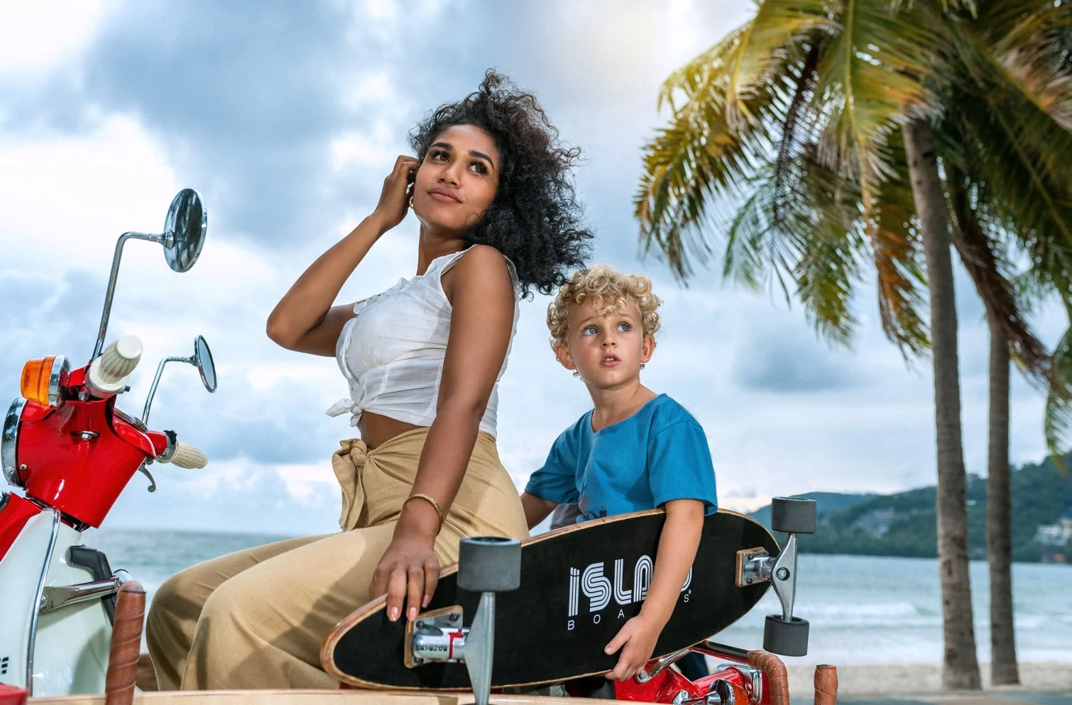 A woman and a young boy on a red scooter near the beach, with palm trees and cloudy sky in the background.
