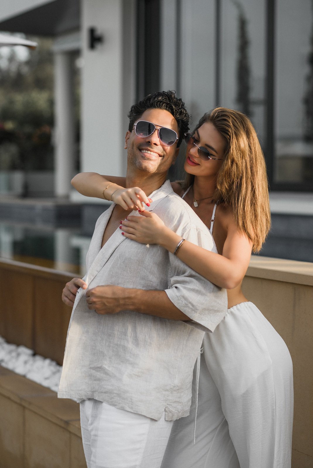A smiling couple wearing sunglasses hugging outdoors near a modern building with large windows.