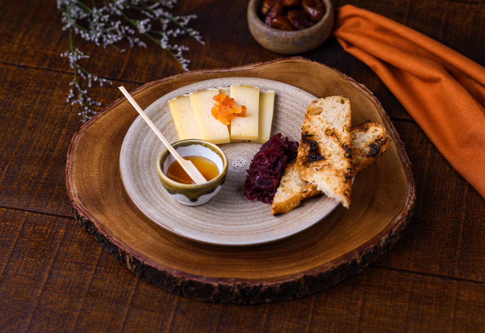 Cheese slices with orange garnish, grilled chicken pieces, red cabbage, honey in a small bowl with a honey dipper, on a wooden plate, surrounded by a flower sprig, a small bowl of dates, an orange napkin, and a wooden table.