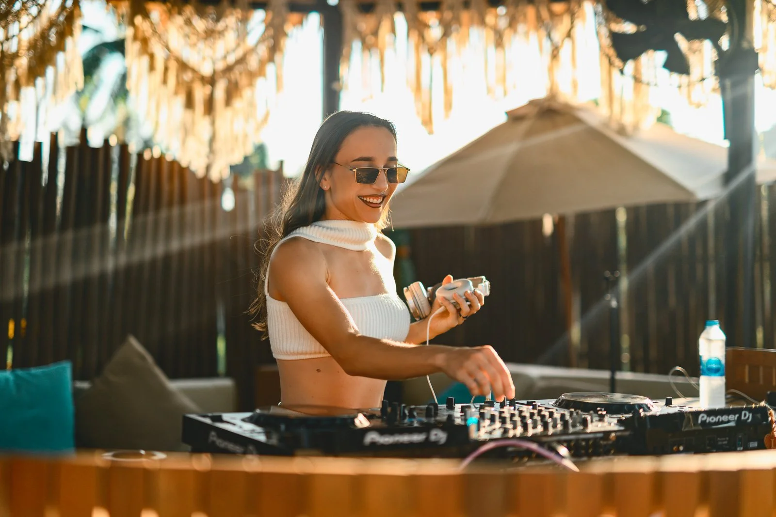 A woman in sunglasses, a white sleeveless top, and long hair, standing outdoors, smiling and DJing with a Pioneer DJ equipment, with sunlight and tropical decor in the background.