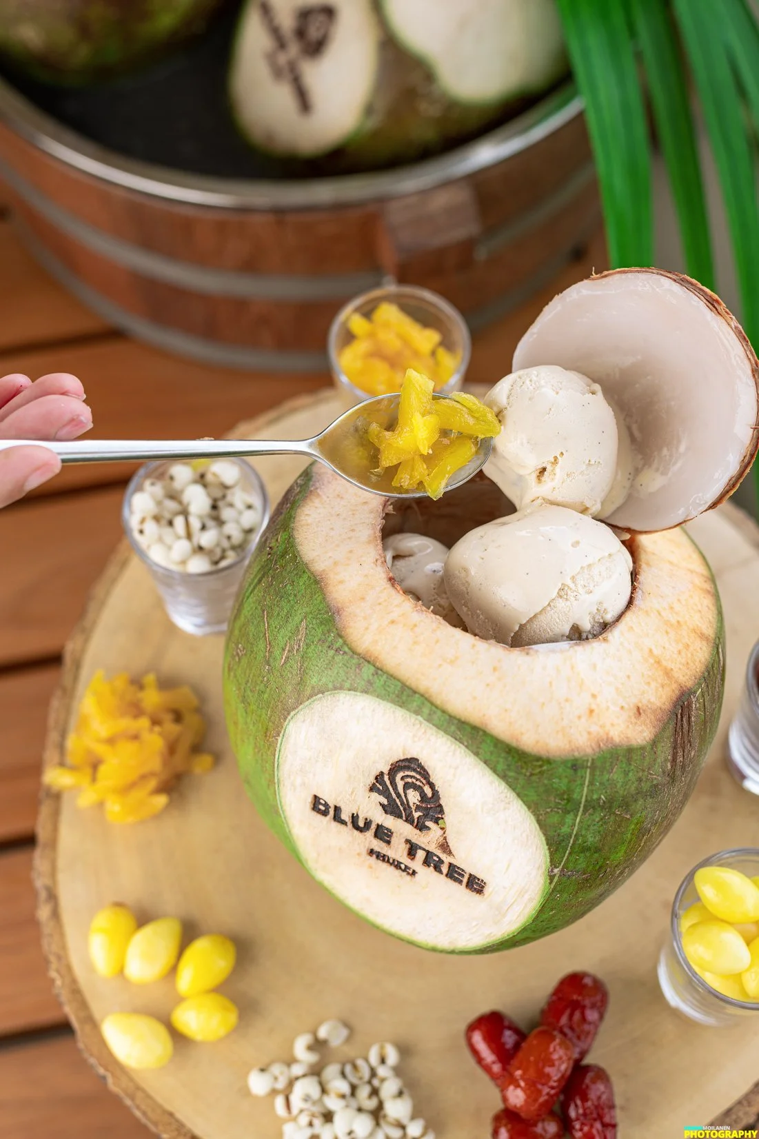 A coconut with its top cut off, filled with ice cream, surrounded by small bowls of yellow pickled peppers, white seeds, and red fruit, on a wooden board.