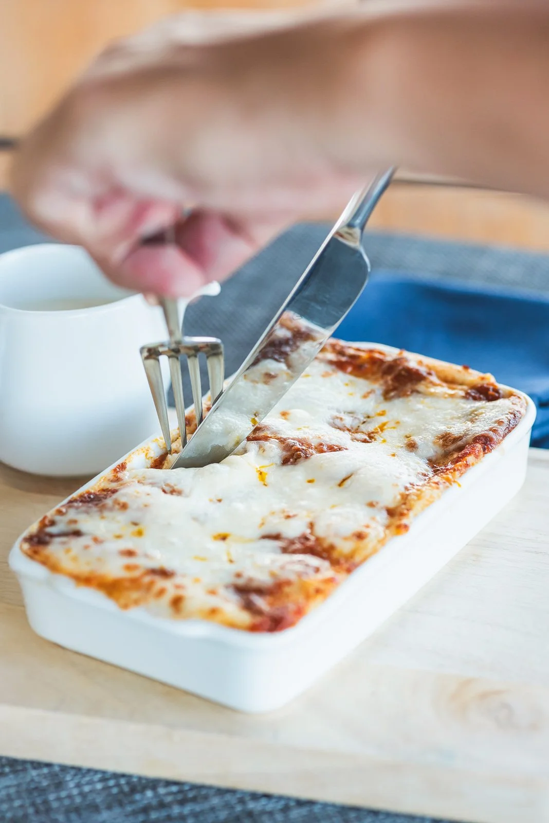Person cutting and serving a portion of baked cheese lasagna with melted cheese on top, on a white dish. A cup of coffee or tea is in the background.