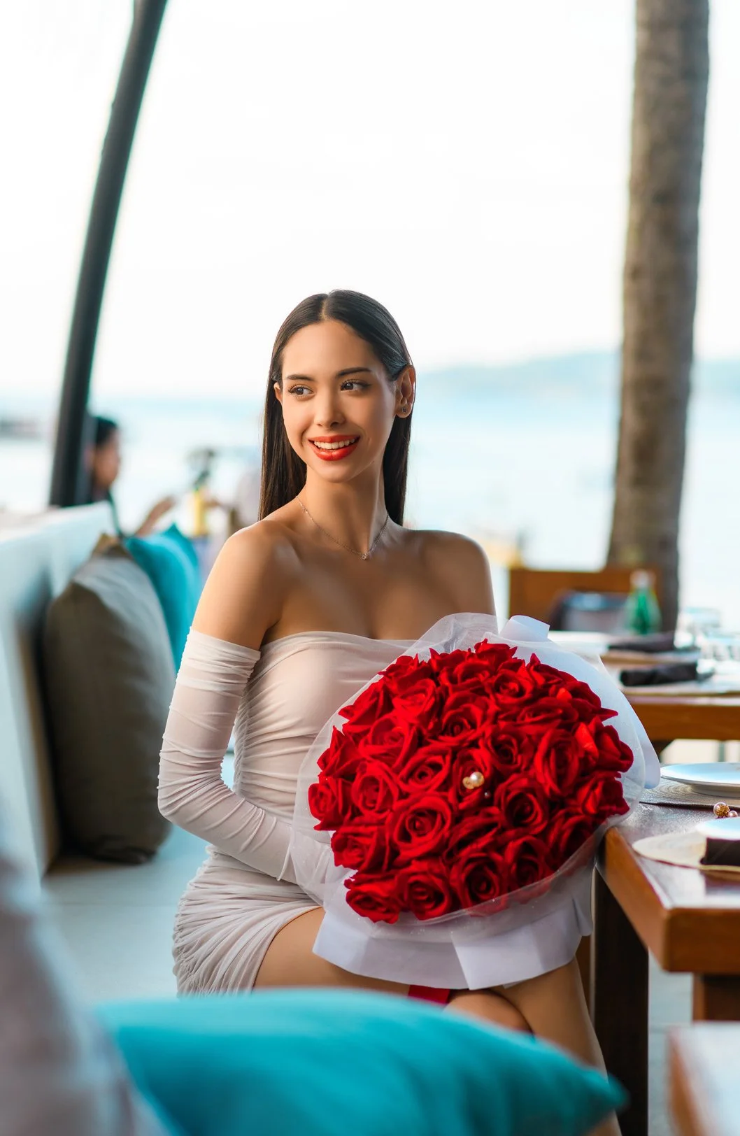 A woman sitting indoors at a restaurant or cafe holding a large bouquet of red roses, with the ocean and a palm tree visible in the background.