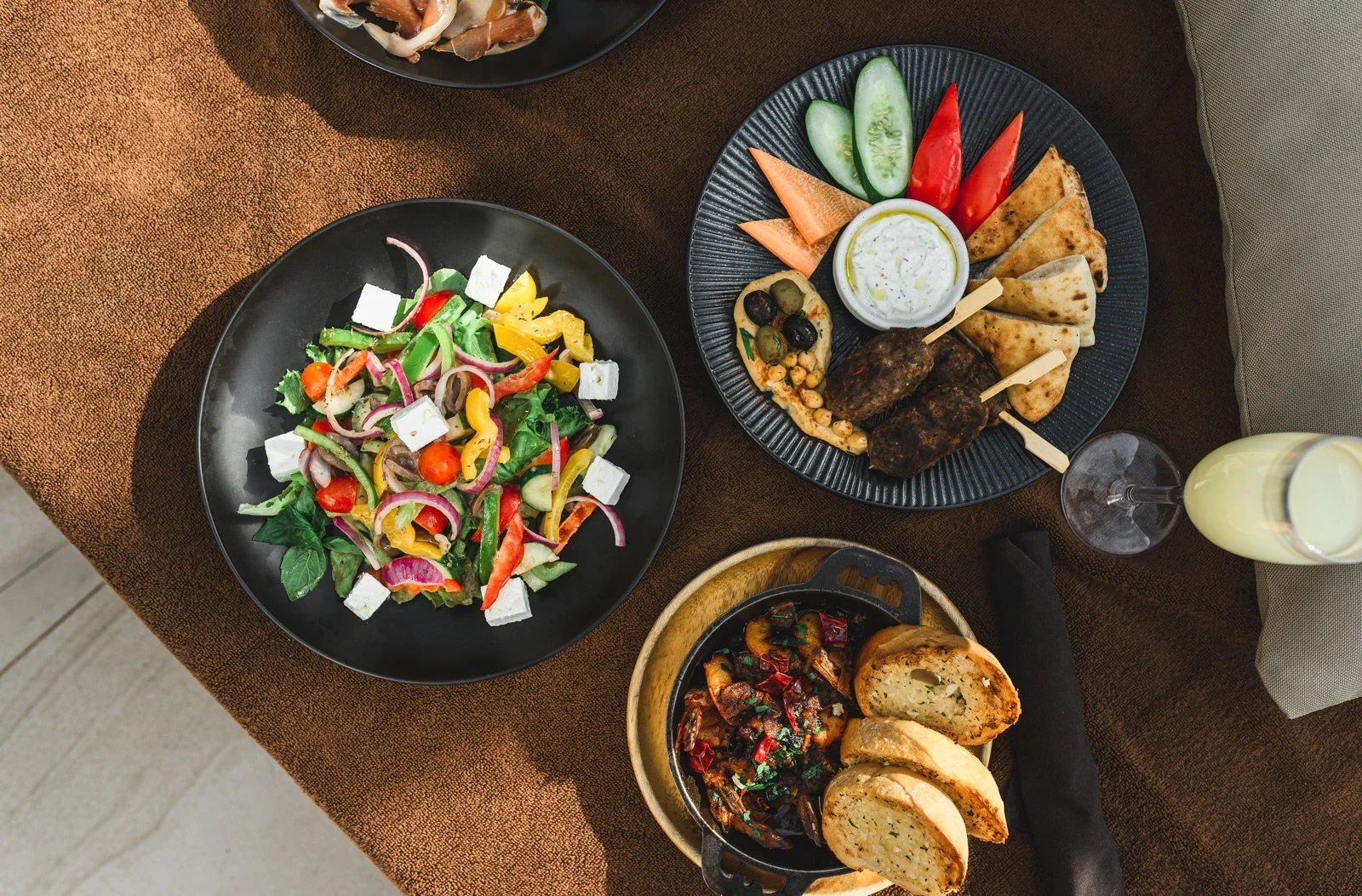 Assorted Mediterranean dishes including a fresh salad with tomatoes, cucumbers, red onions, olives, feta cheese, and greens; a platter with grilled meat, pita bread, vegetables, and dipping sauces; a bowl with eggplant dish, and sliced garlic bread; 