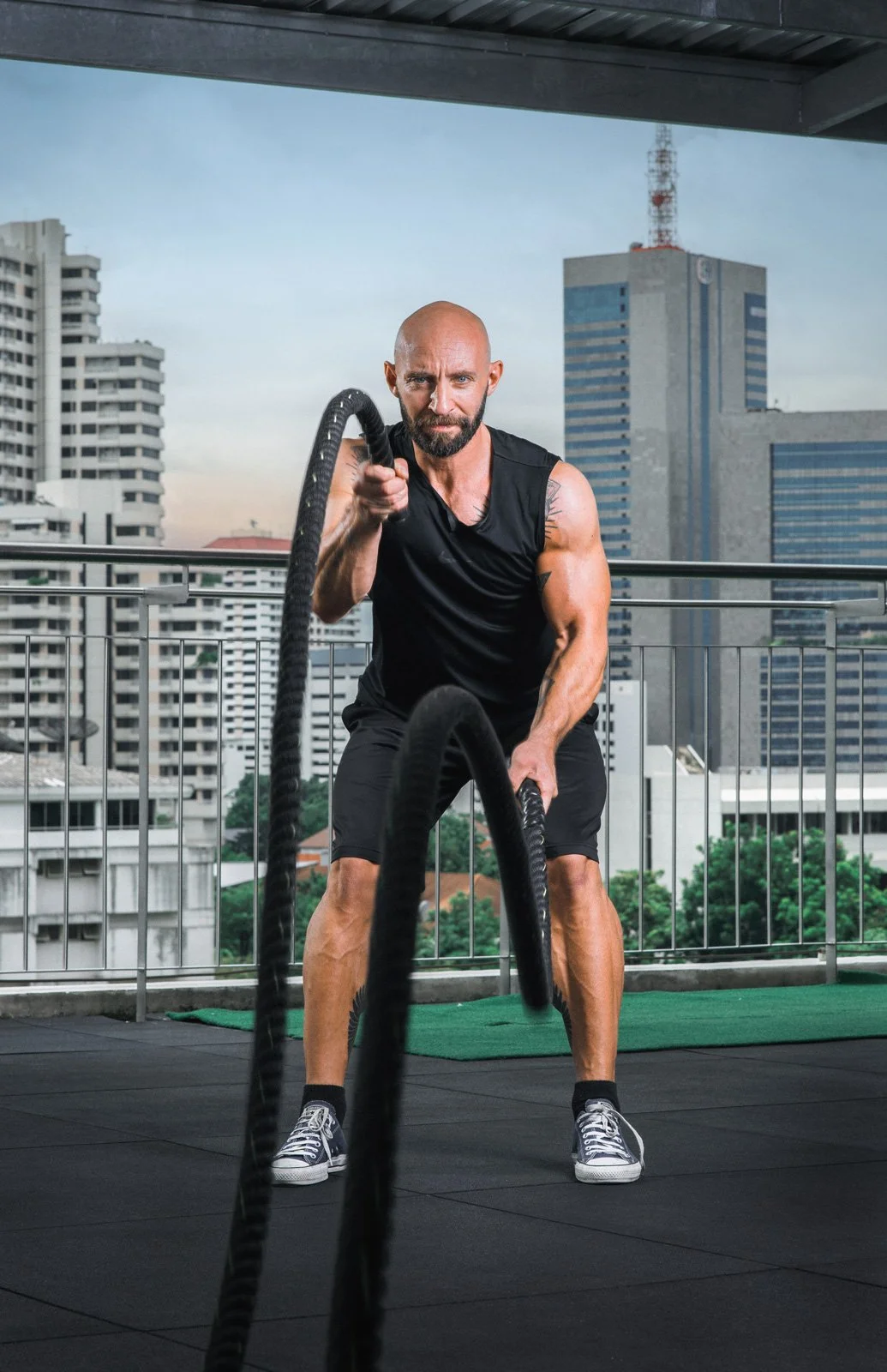 A man with a beard, wearing a black sleeveless shirt and shorts, is exercising with battle ropes on a rooftop gym with a cityscape background.
