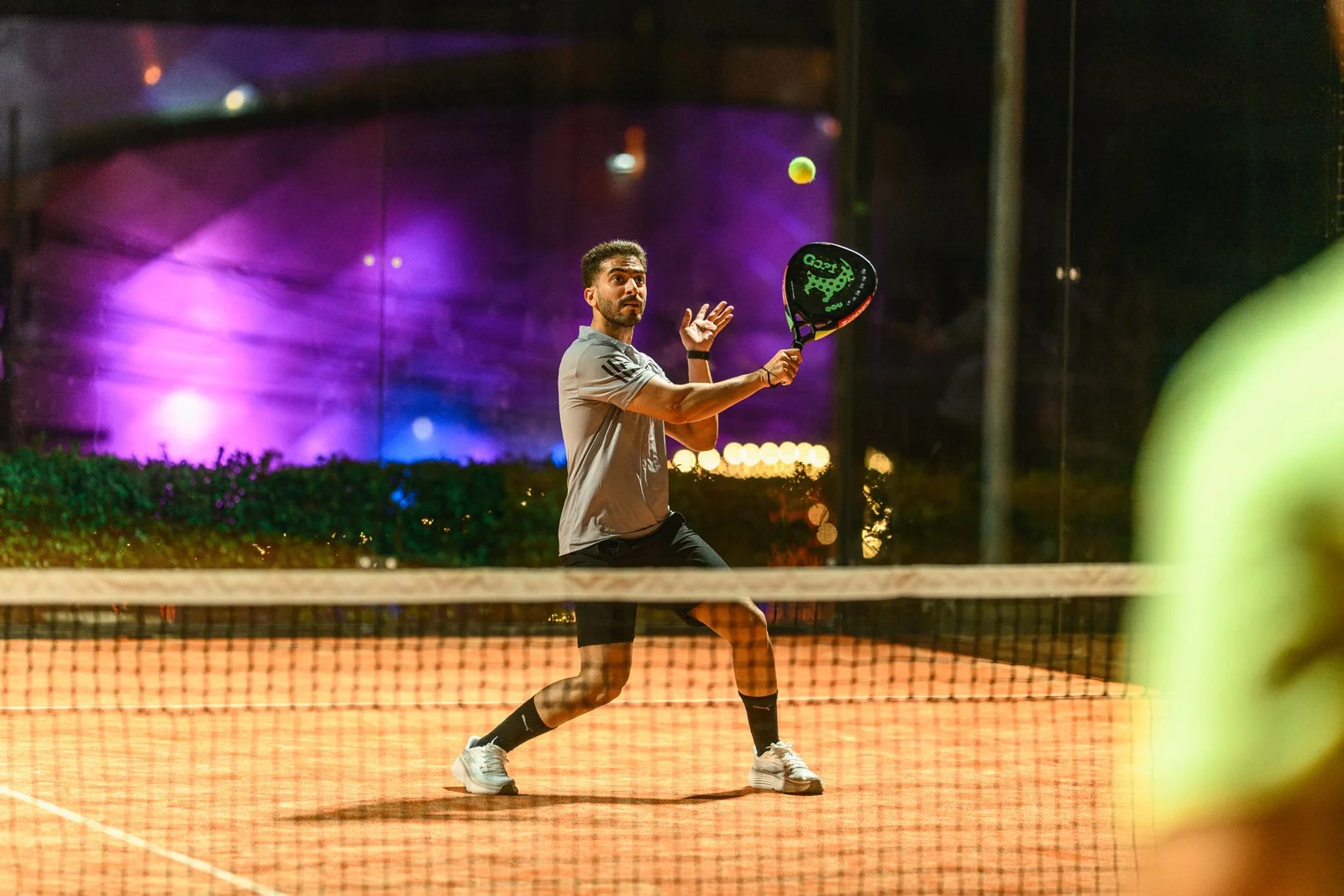 A man playing padel on an outdoor court at night, preparing to hit a tennis ball with a racquet, with colorful purple and pink lighting in the background.