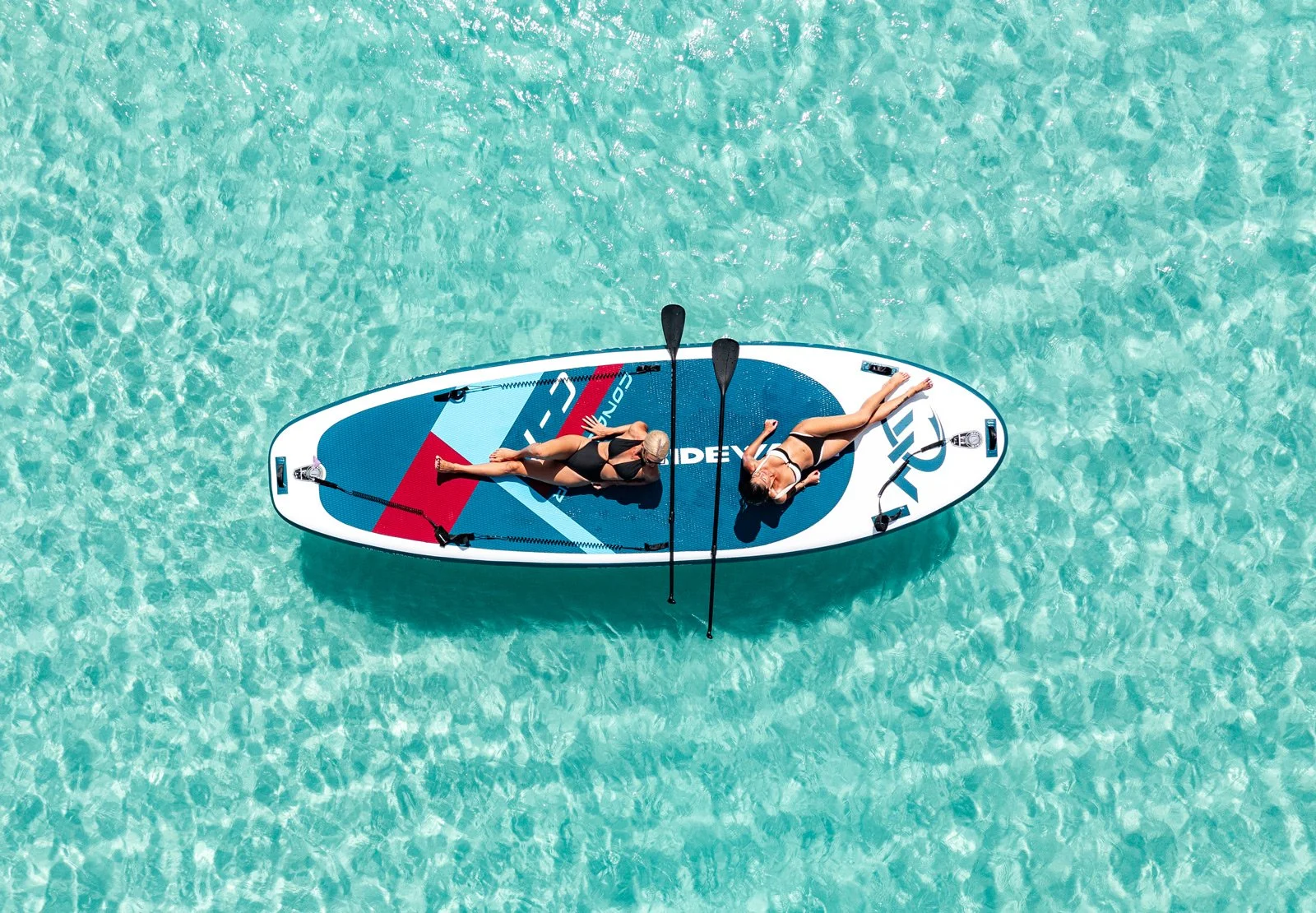 Two women in swimsuits relaxing on a paddleboard in shallow, clear turquoise water.