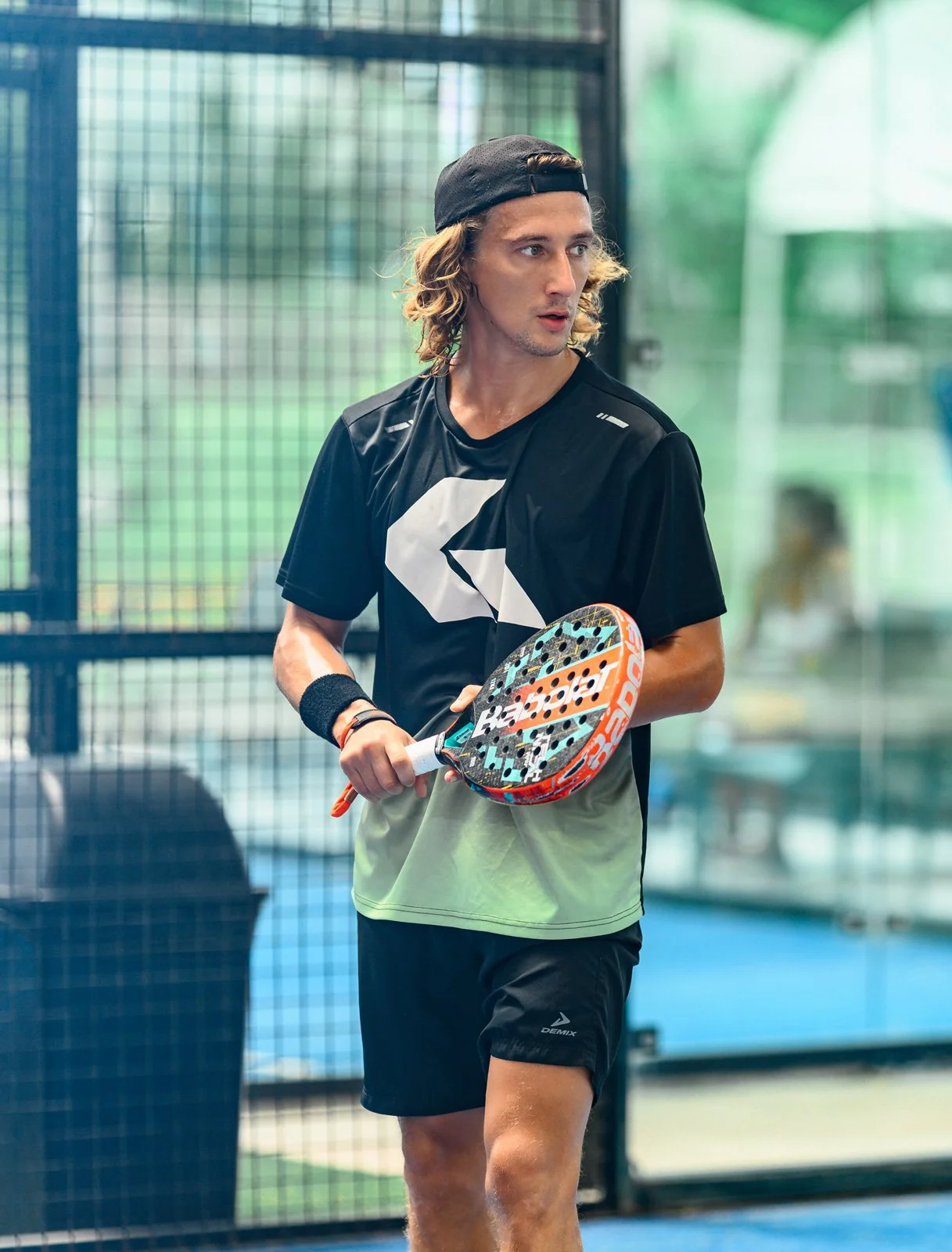 A young man with long curly hair, wearing a black cap backwards, black and green athletic shirt, and black shorts, holding a padel racket in an indoor padel court.