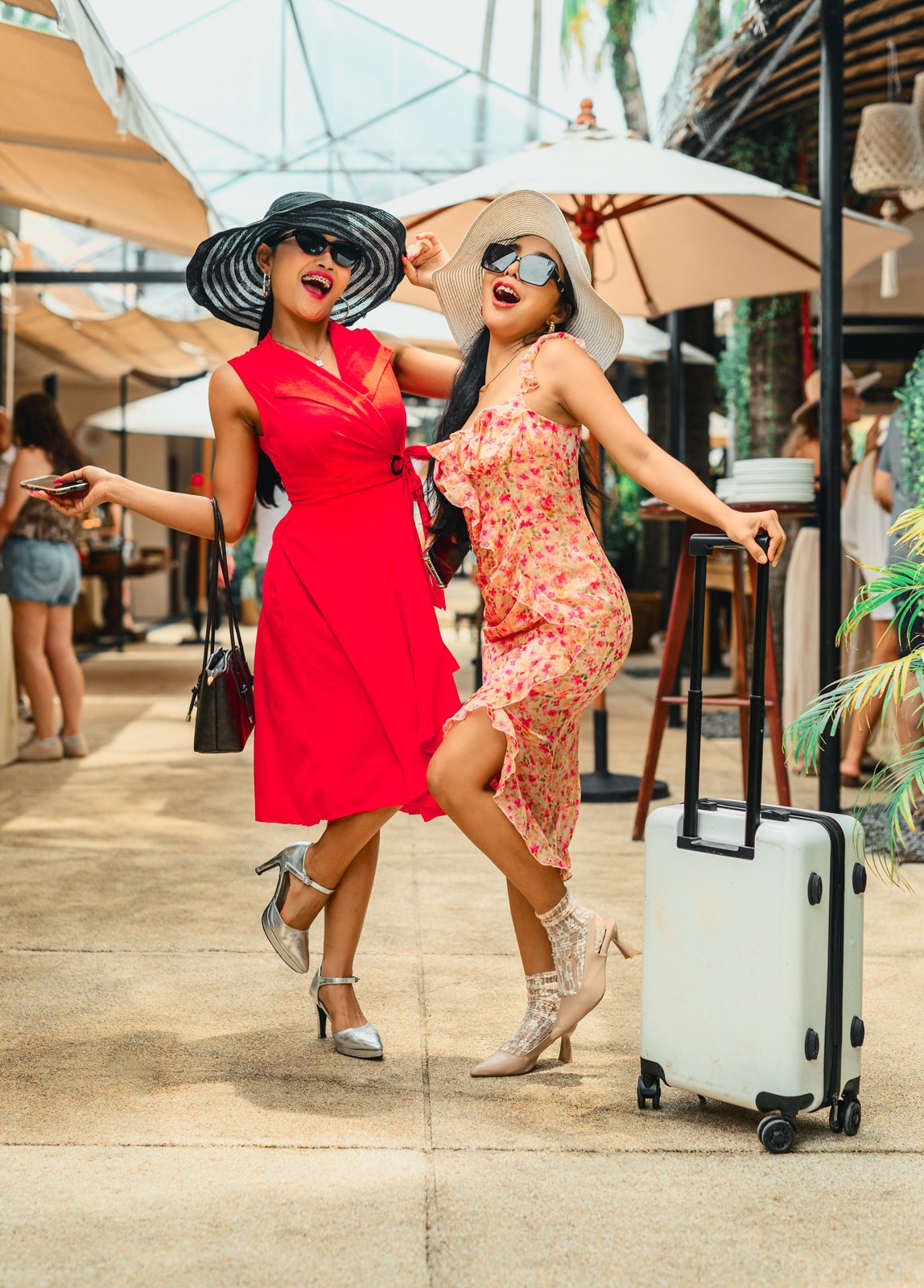 Two women in colorful dresses and large sun hats posing happily at an outdoor venue, with one holding a suitcase.