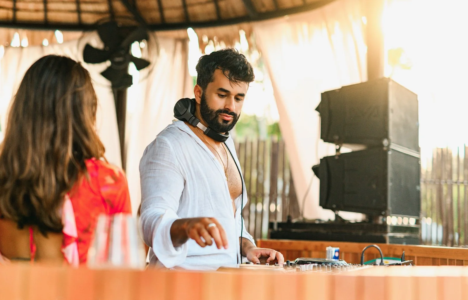 A man with dark hair and beard, wearing a white shirt and a gold chain, is DJing at an outdoor event. He has headphones around his neck and is adjusting music equipment on a table. A woman with brown hair and a red floral top faces him. The backgroun