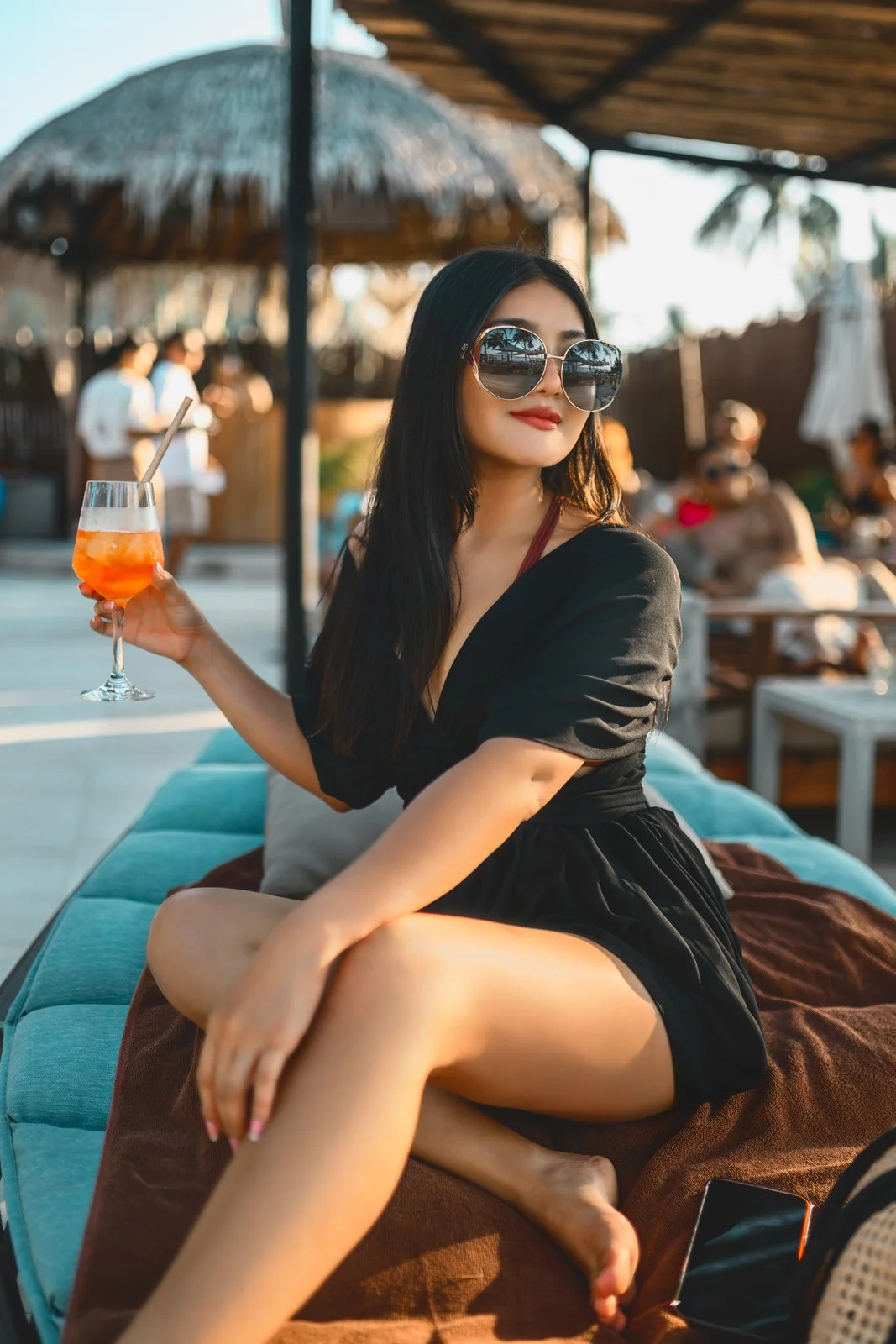 A woman with long dark hair and sunglasses sitting outdoors at a bar or poolside, holding a colorful cocktail and wearing a black dress. There are other people in the background and a thatched umbrella.