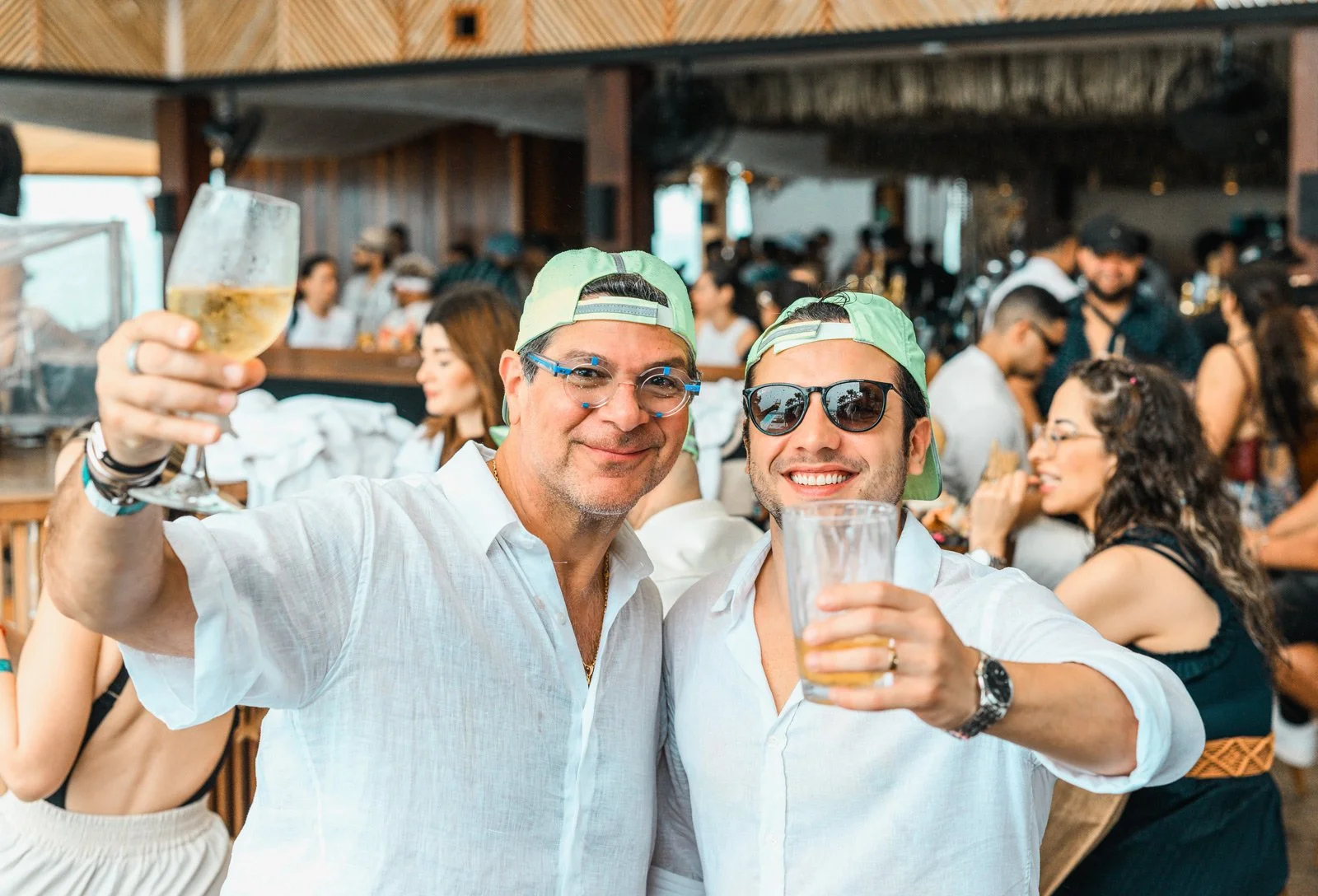 Two men in white shirts wearing green baseball caps and sunglasses, smiling and holding drinks, at a lively celebration or party in an indoor venue with many people in the background.