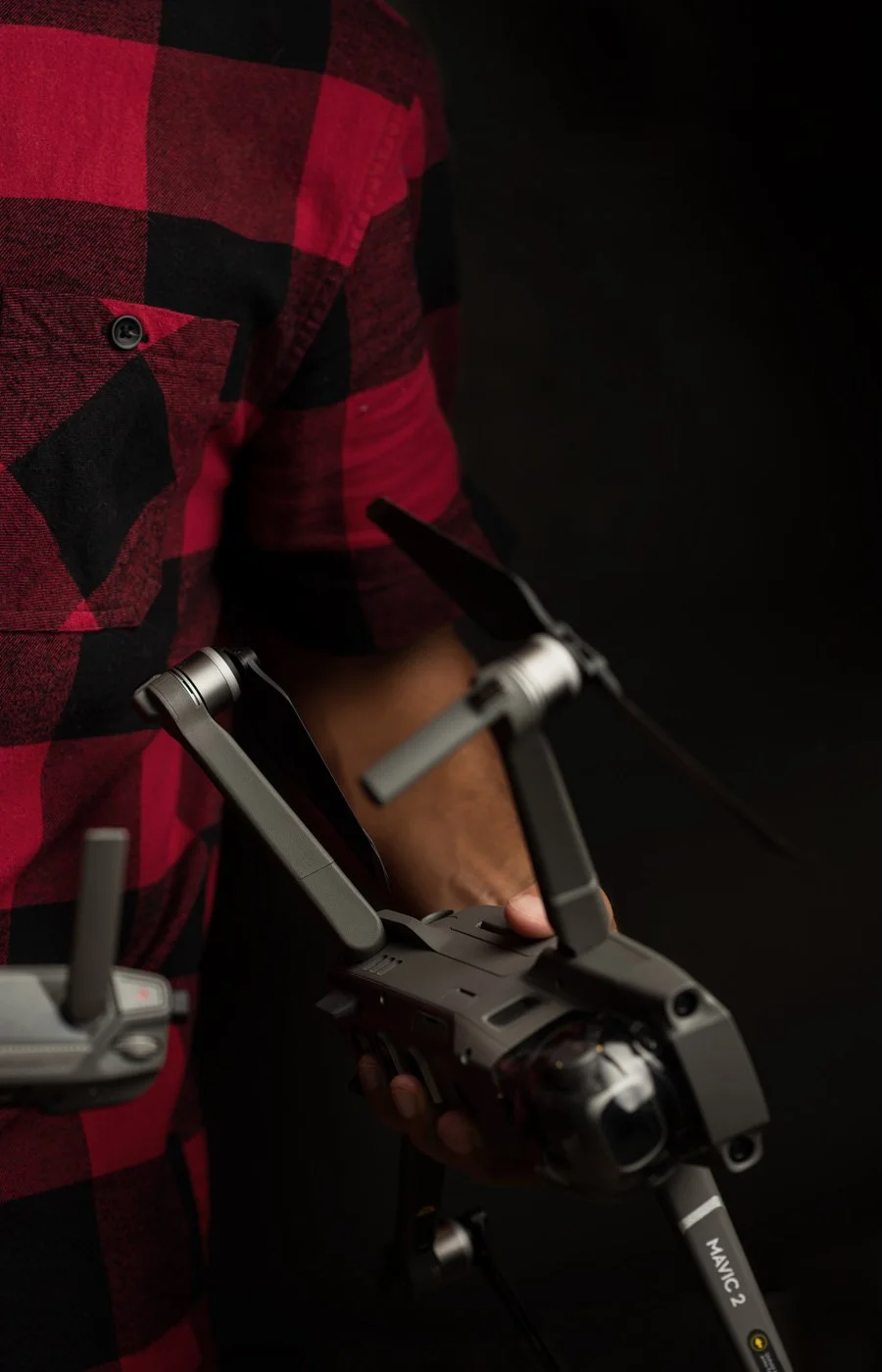 A person in a red and black checkered shirt holding a drone with a camera and remote control against a dark background.