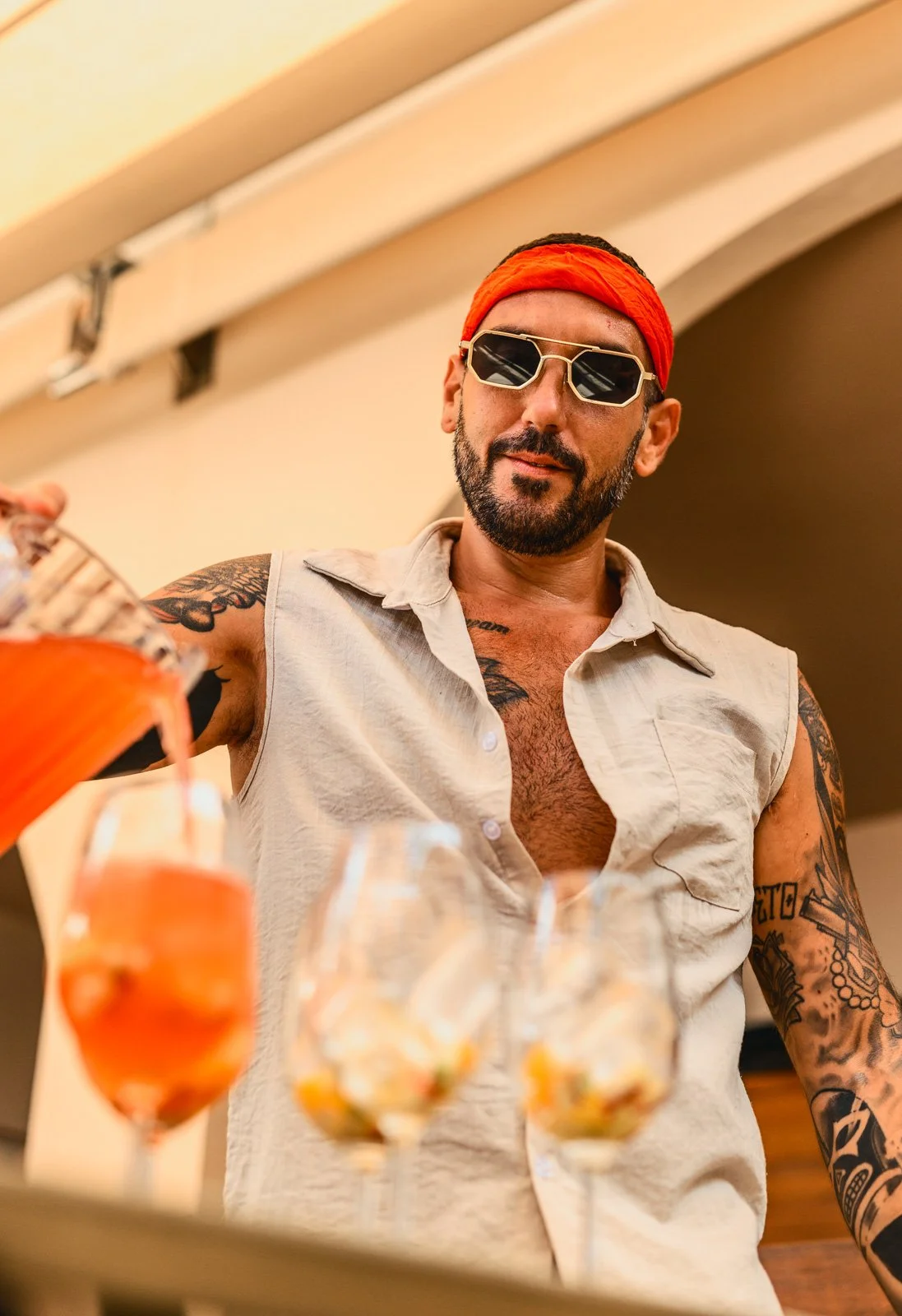 A man with tattoos, wearing sunglasses, a red headband, and a sleeveless shirt, pouring drinks at a party or gathering.