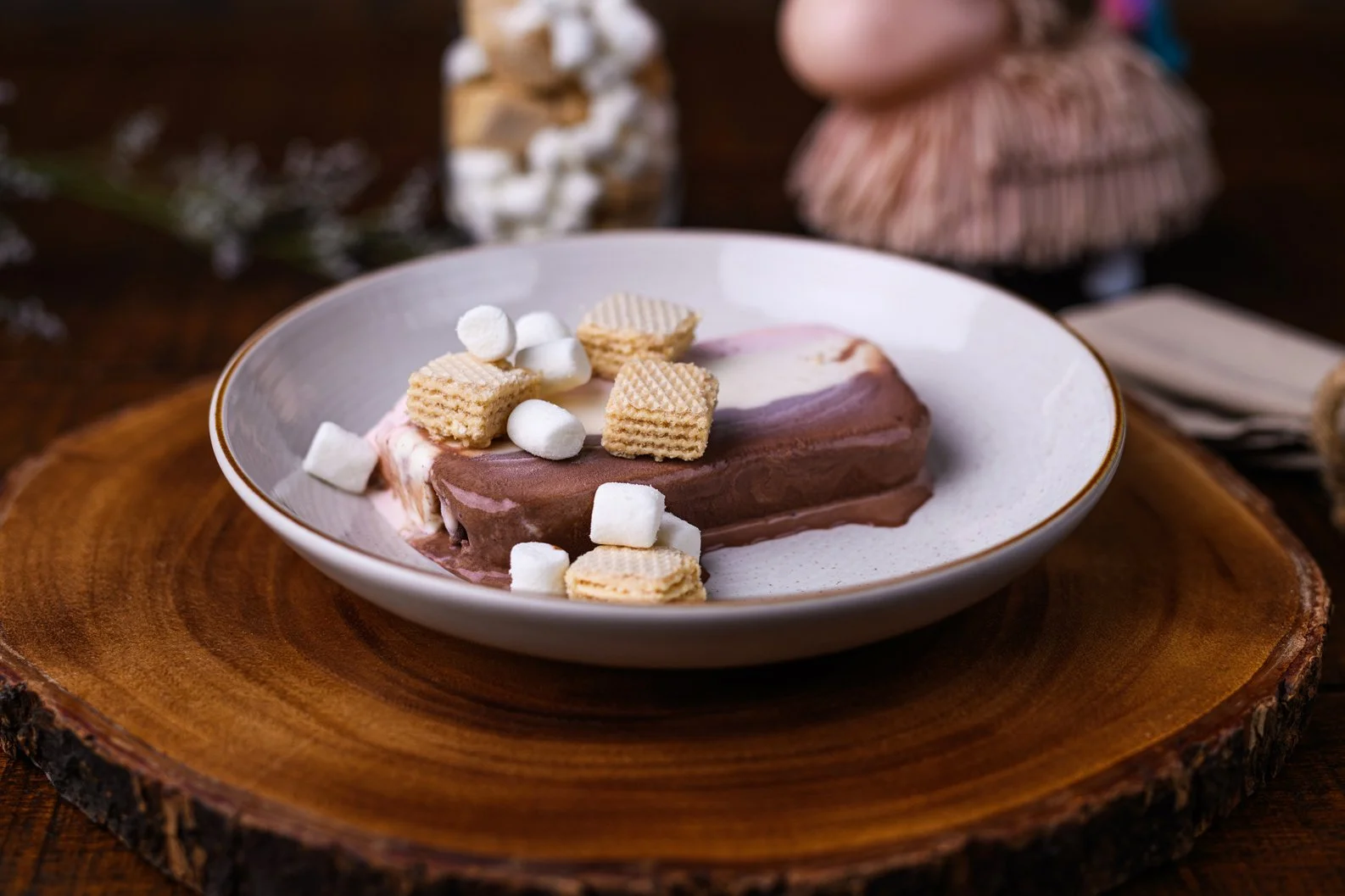 Chocolate ice cream topped with mini marshmallows and wafer cookies on a white plate, placed on a wooden serving board.