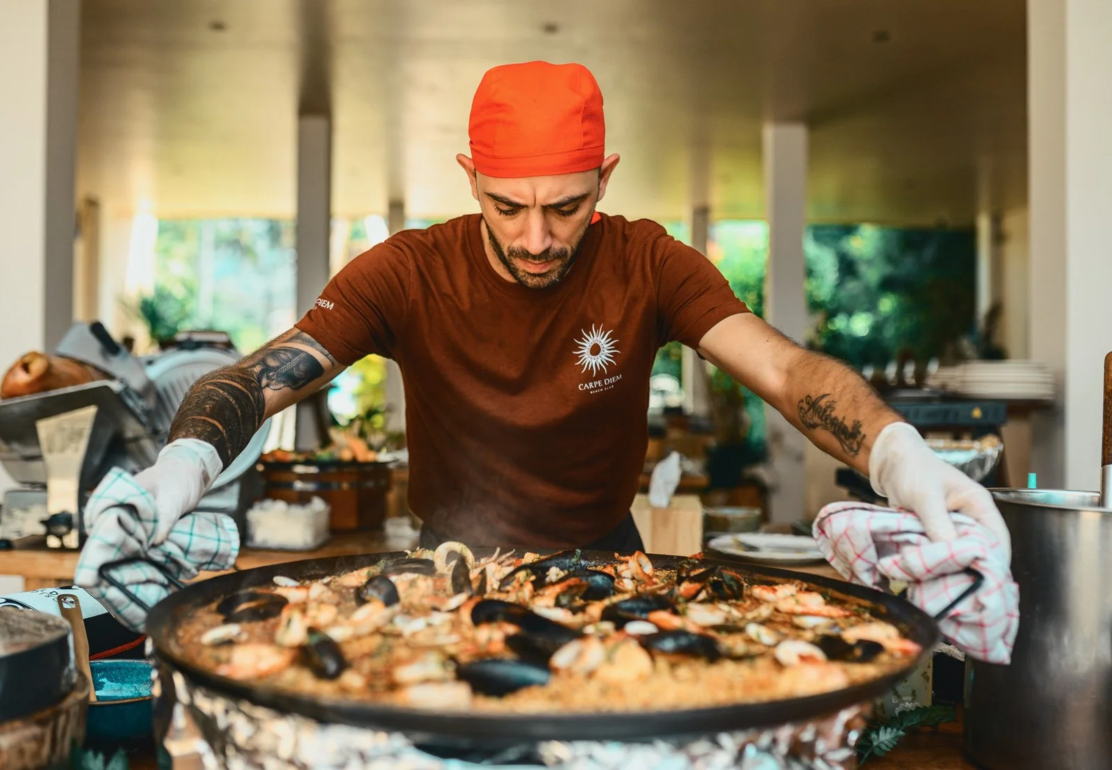 Man cooking seafood paella in a large pan at a restaurant or outdoor venue, wearing a red head covering and brown shirt with a sun logo.