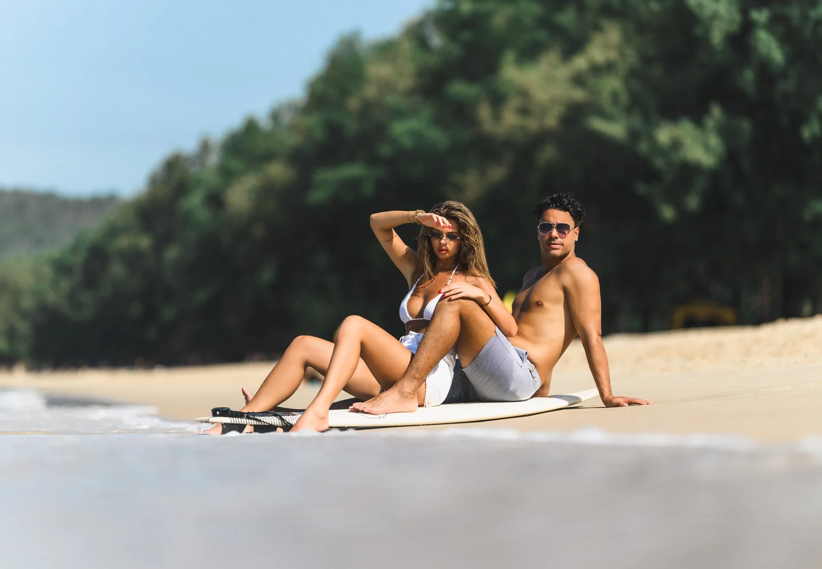 A man and woman in swimwear sitting on a surfboard on the beach, with trees in the background.
