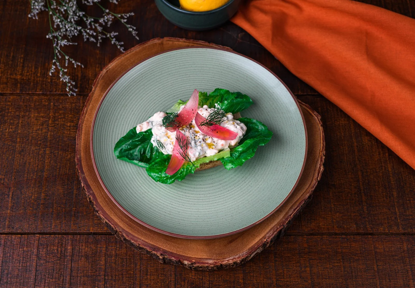 Open-faced sandwich with lettuce, creamy filling, pickled onions, and herbs on toast, served on a ceramic plate on a wooden surface