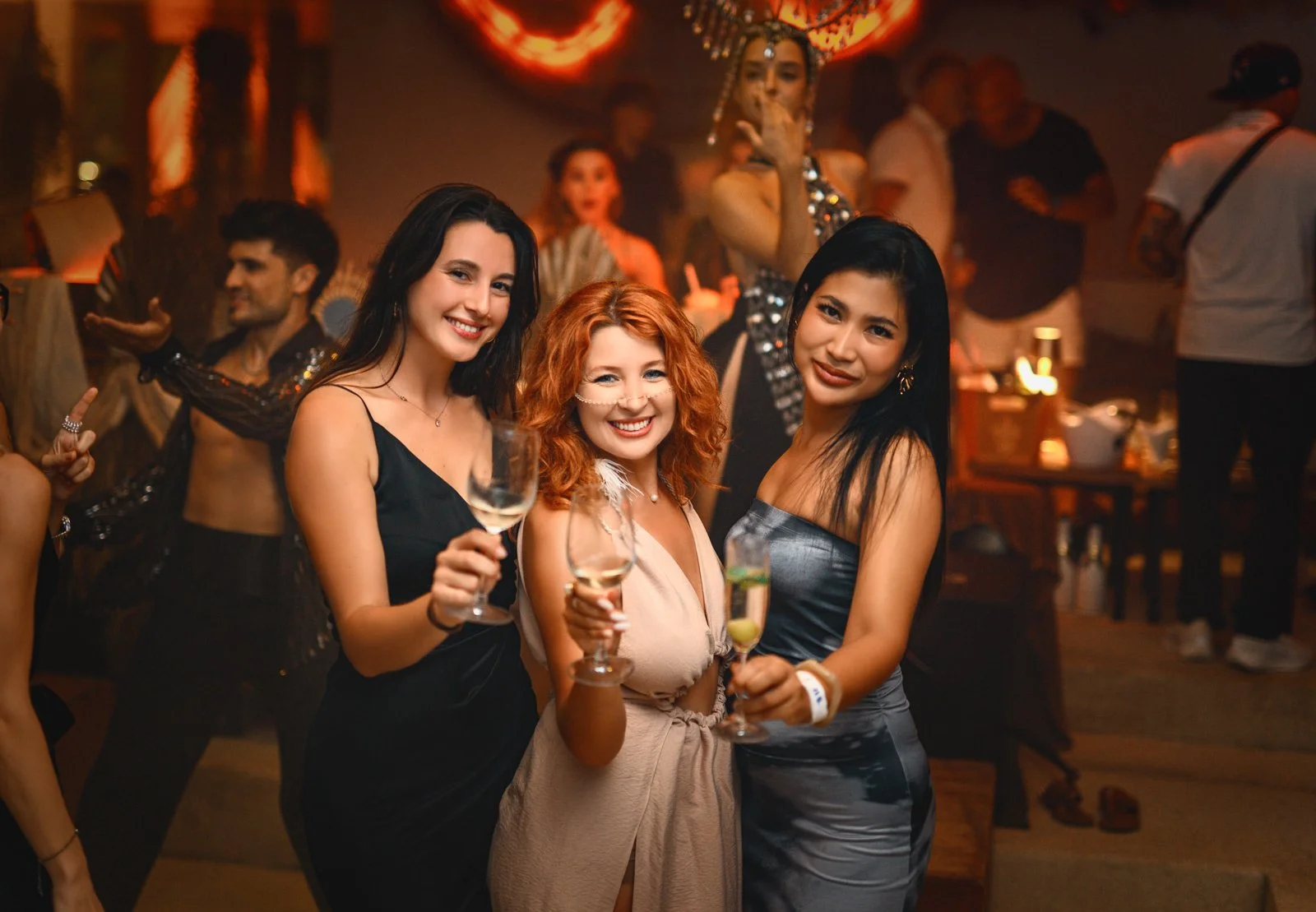 Three women at a party or celebration, smiling and holding wine glasses. The background shows other people dancing and socializing in a dimly lit room.