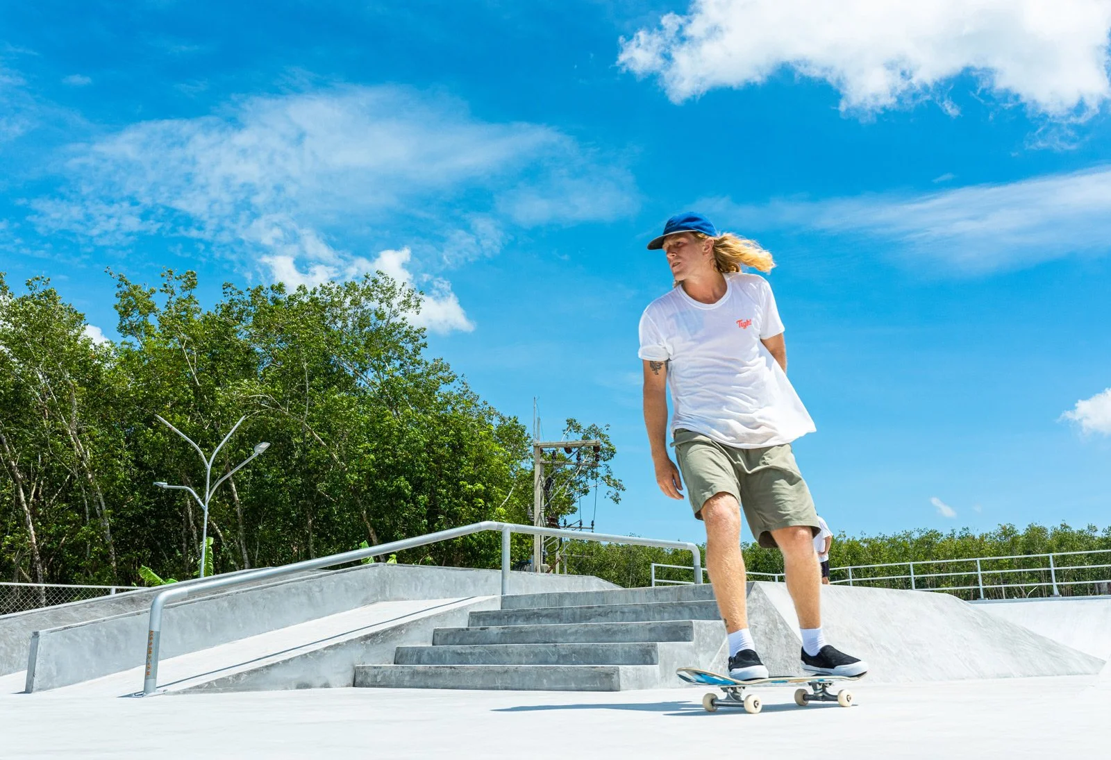 Young woman skateboarding at a skate park on a sunny day with blue sky and green trees in the background.