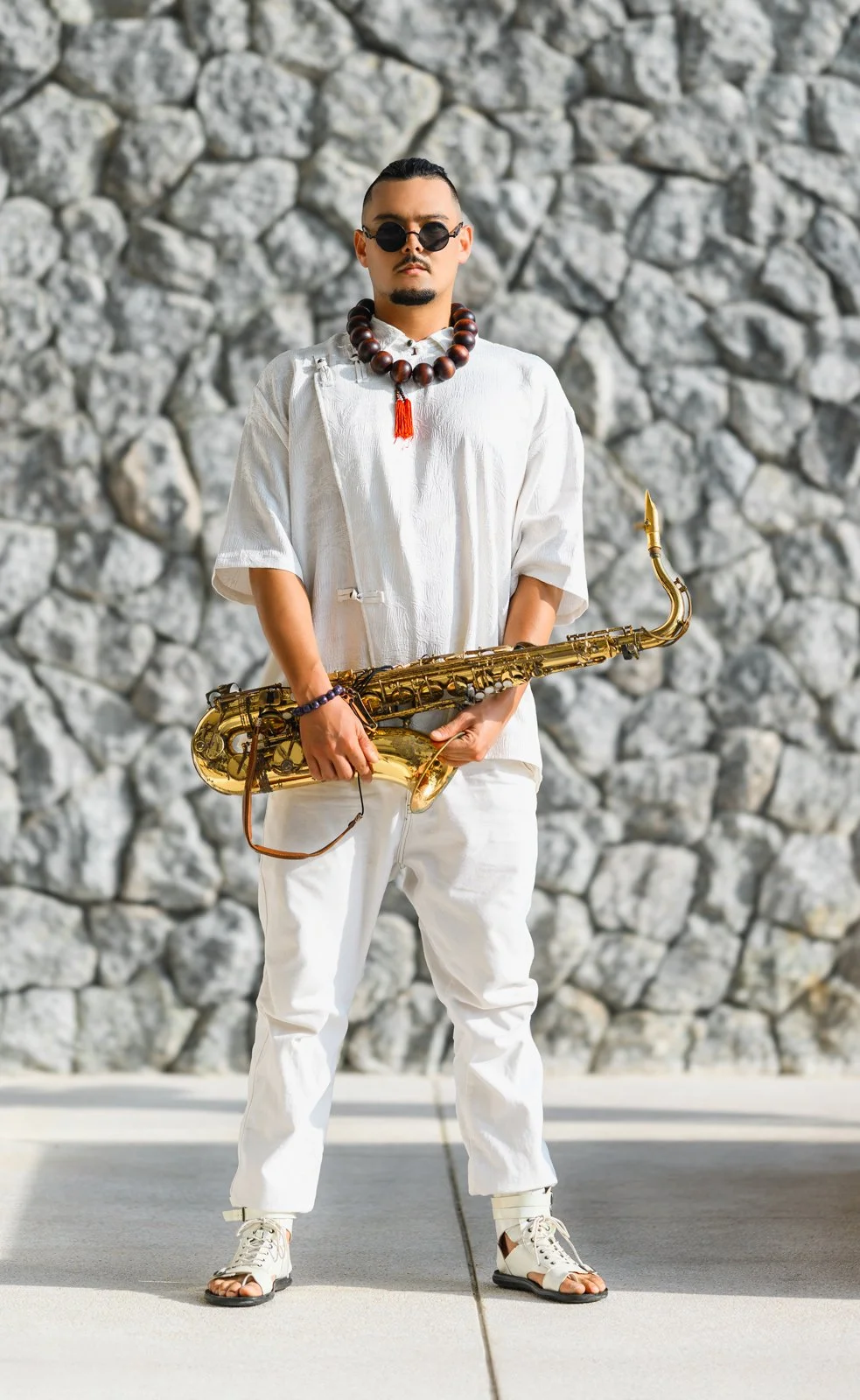 A man in white clothing stands in front of a stone wall, holding a saxophone, wearing sunglasses, a large beaded necklace, and sandals.