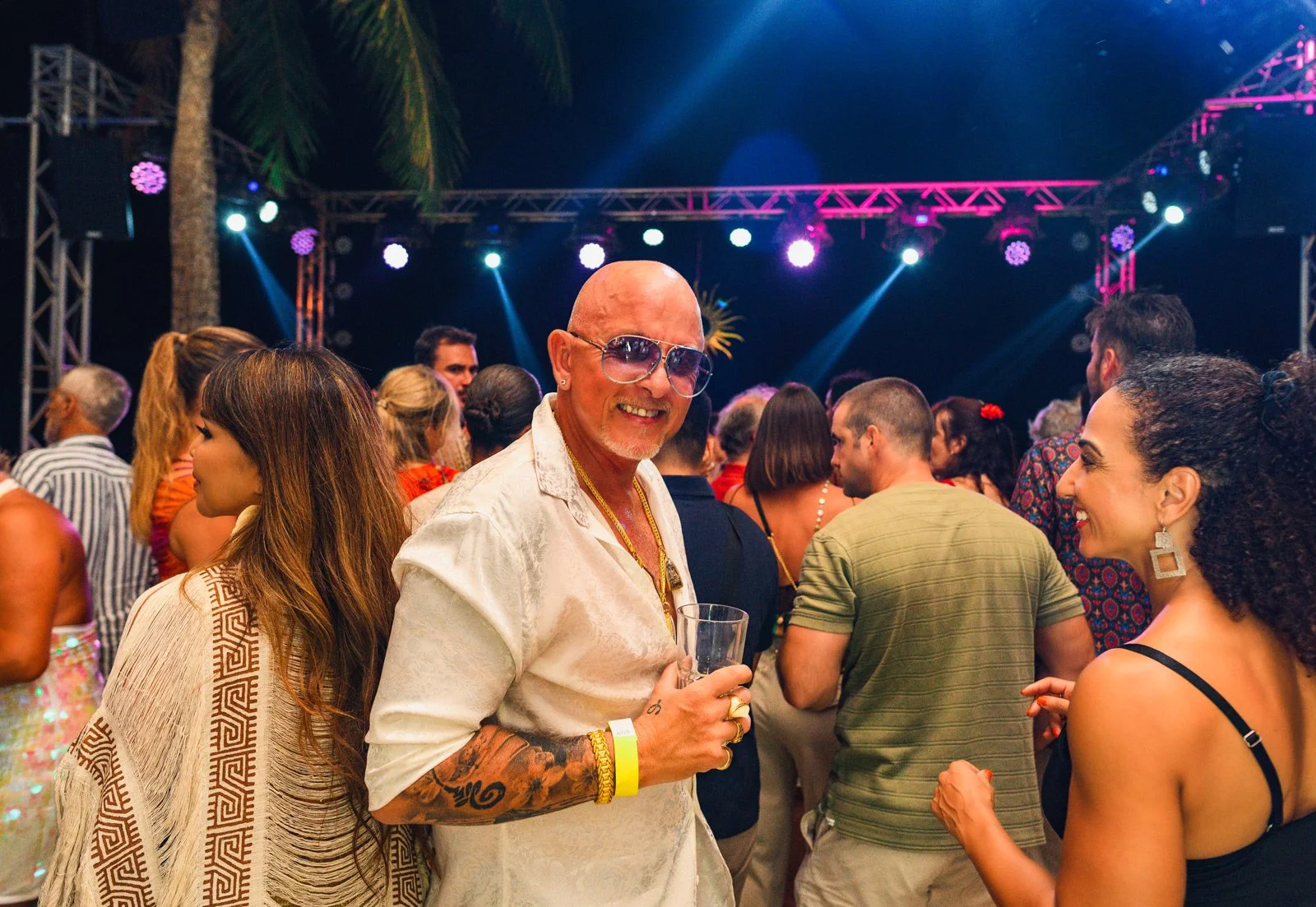 A diverse group of people attending a lively outdoor concert or party at night, with colorful stage lights and a palm tree in the background. The central figure is a bald man with tattoos, wearing sunglasses, smiling while holding a drink, surrounded
