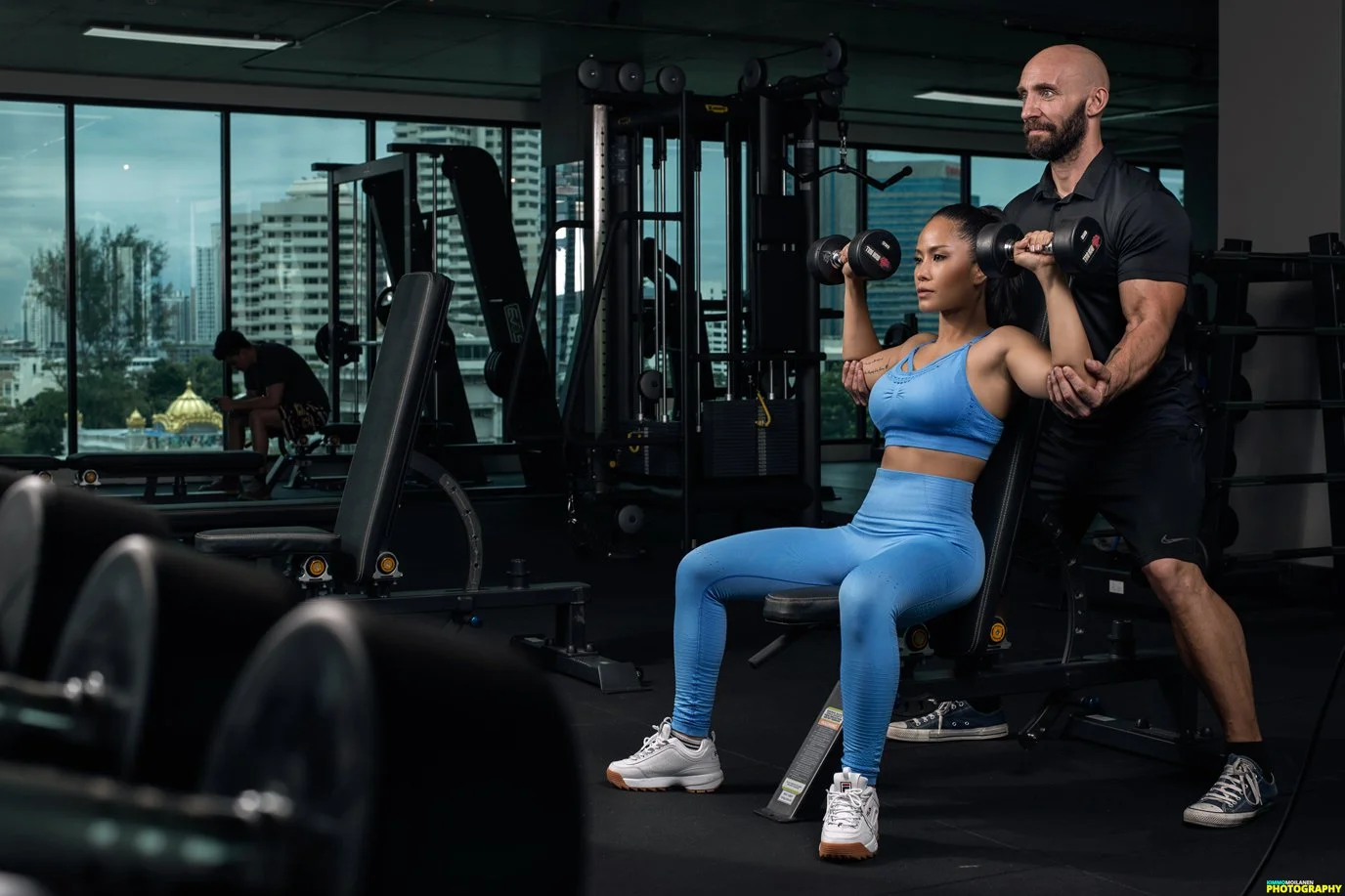 A woman in a blue workout outfit sitting on a chest press machine doing shoulder presses with a trainer standing behind her, guiding her.