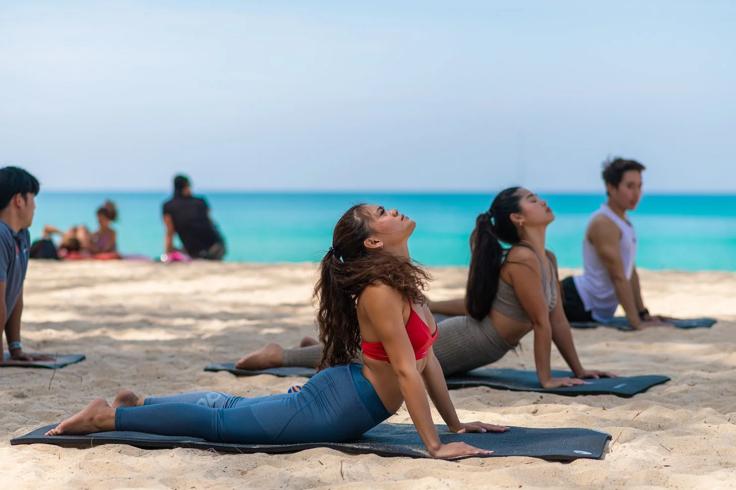 Group of people practicing yoga underwater on a sandy beach, with ocean and blue sky in the background.