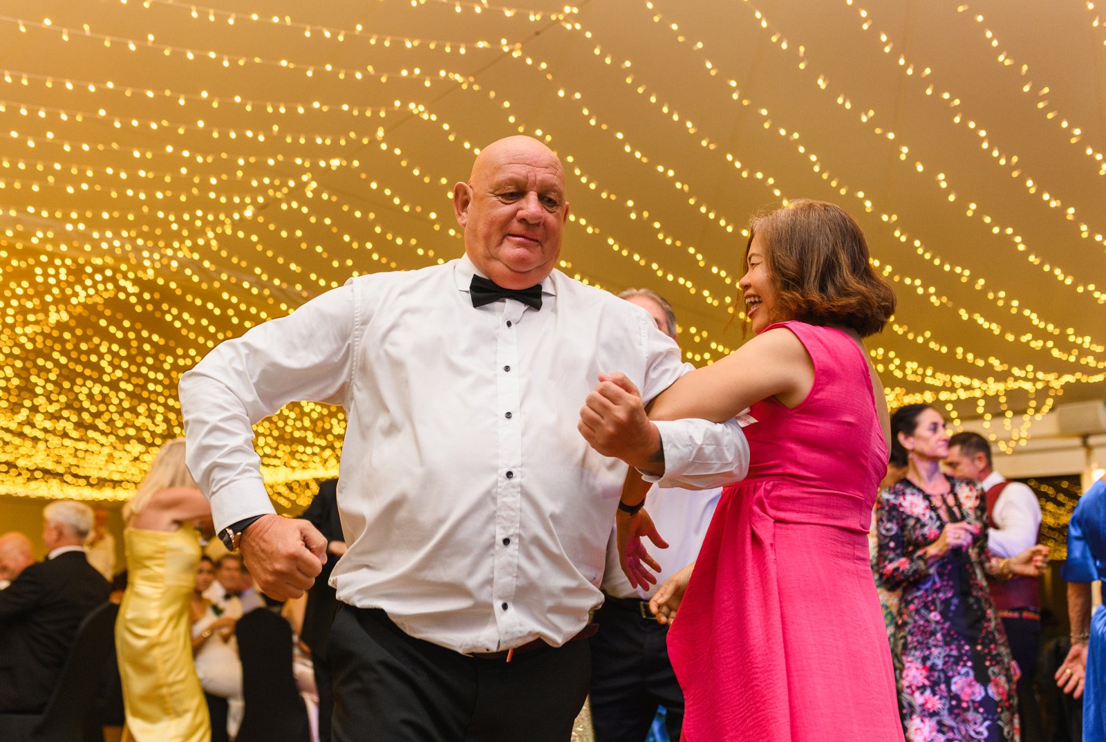 A man and woman dancing and smiling at a festive event with string lights overhead, surrounded by other guests in formal attire.