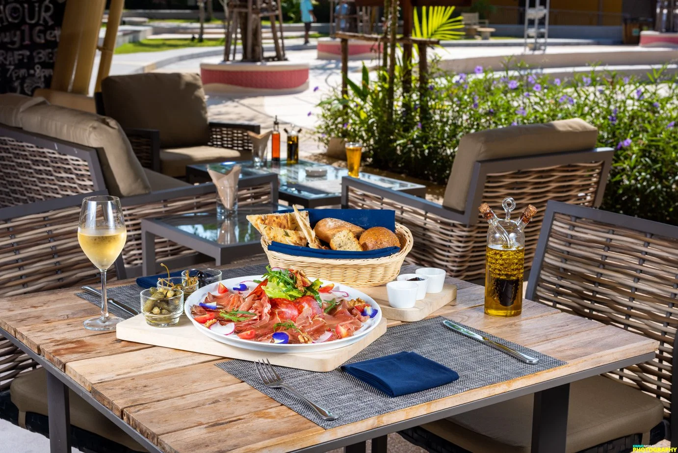 Outdoor restaurant table set with a cheese and bread basket, a plate of prosciutto and greens, a glass of white wine, a bottle of olive oil, cup of salt, and napkin, with chairs around and potted plants in the background.