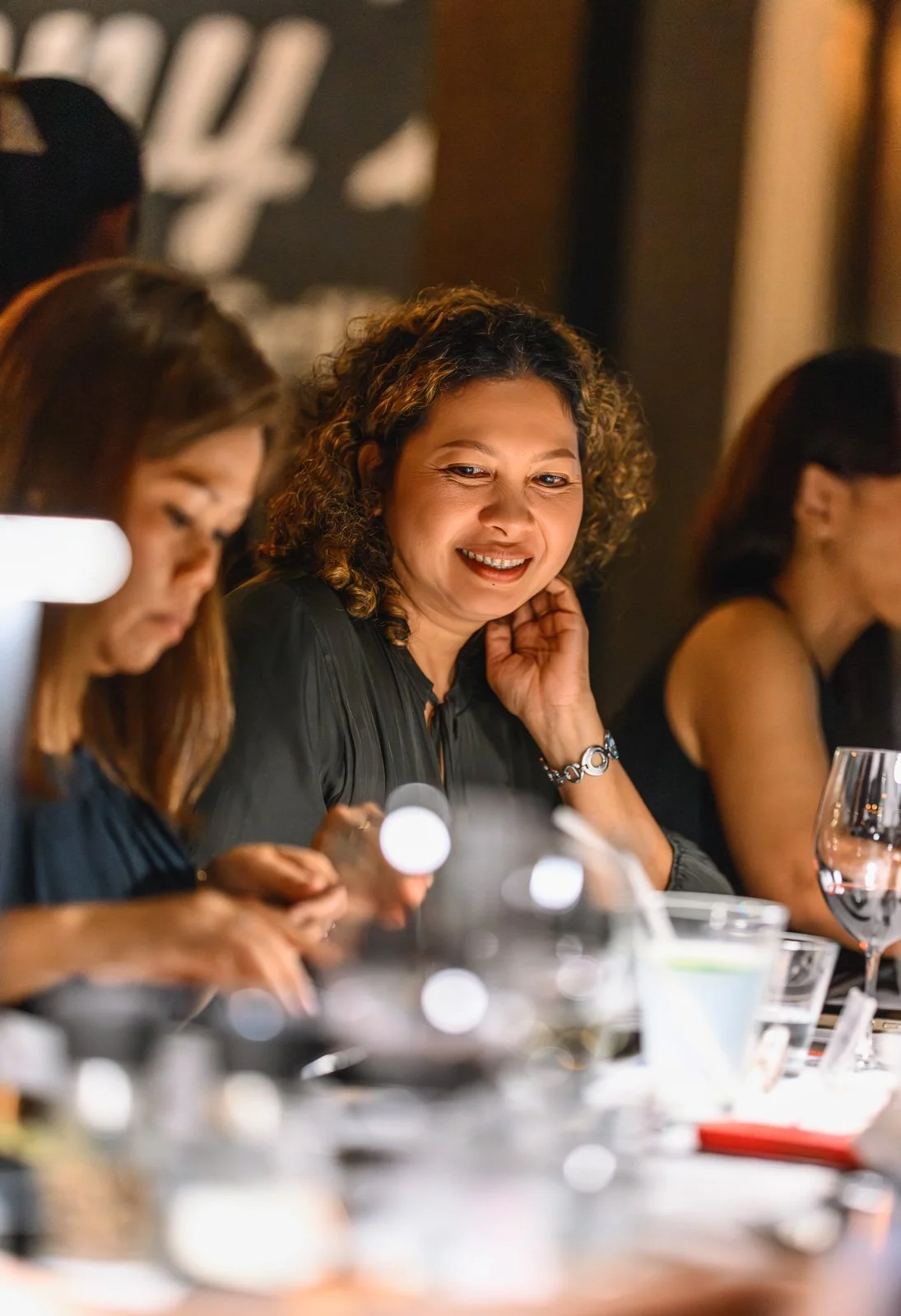 A group of people sitting at a dinner table, with one woman smiling and looking toward the camera, while others focus on their food or drinks.