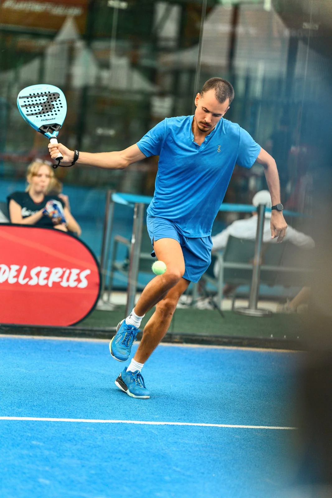 Male padel player in blue attire hitting a ball on an indoor court, with a woman in the background watching.