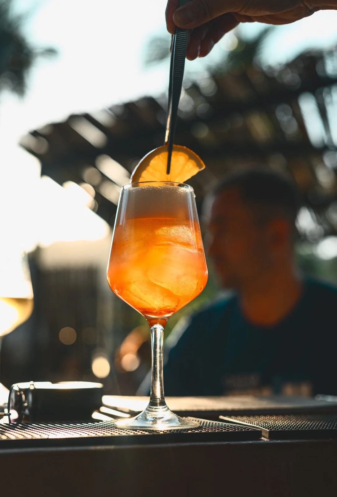 A colorful cocktail with orange slices and ice cubes in a tall stemmed glass on a bar counter, with a blurry person in the background and outdoor setting.
