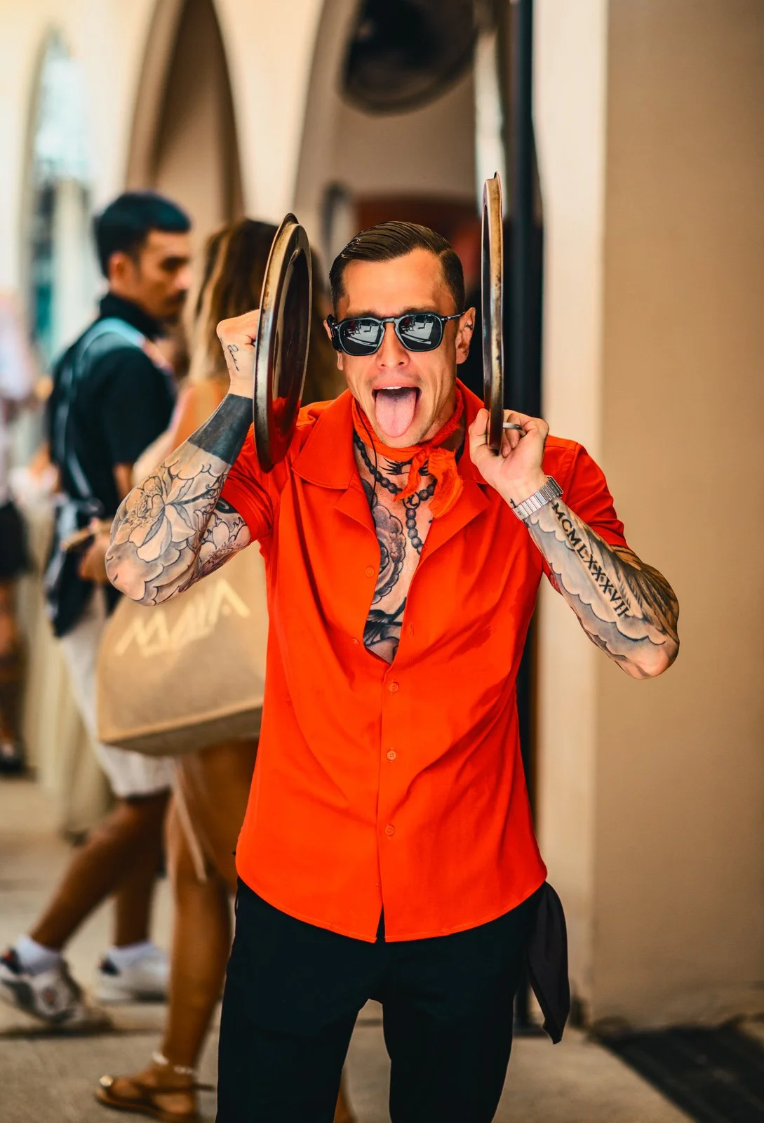Man wearing sunglasses and bright orange shirt, sticking out his tongue, holding two frying pans on his head, tattoos on arms and chest, tattoos with Roman numerals on right arm, tattooed neck, in a shopping mall.