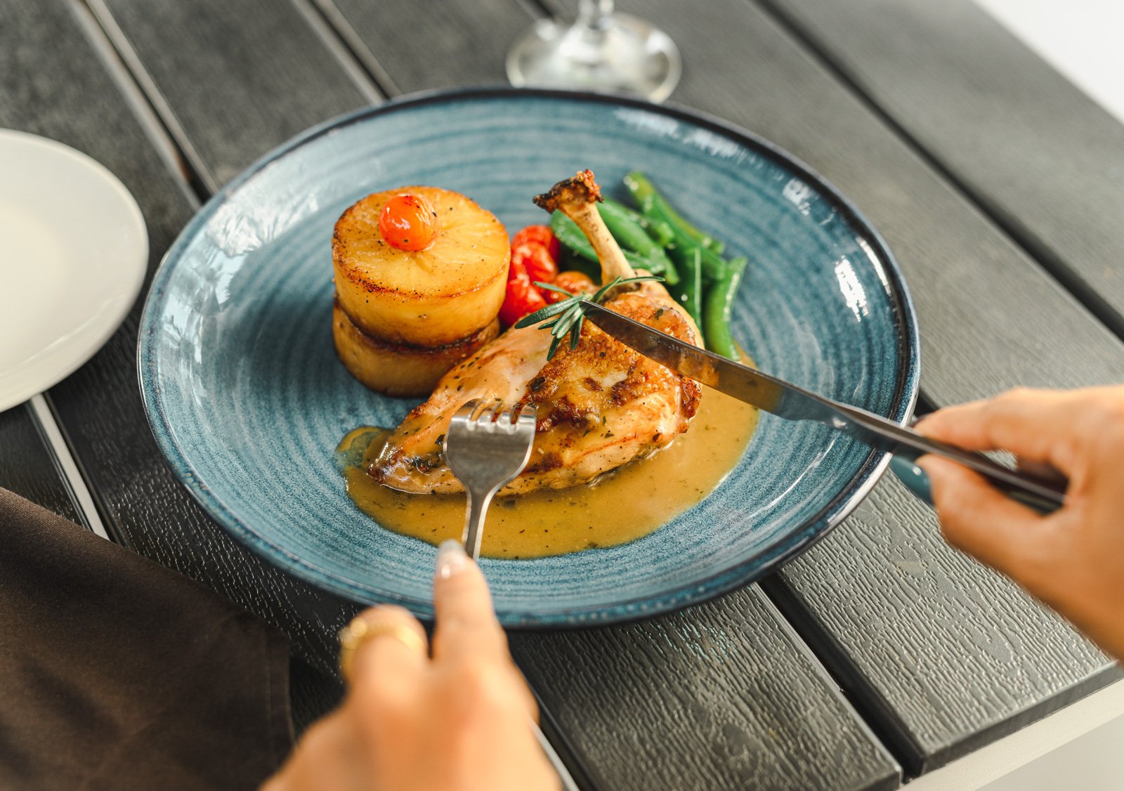 A person cutting into a cooked chicken leg with a sauce, served with a roasted potato topped with a cherry tomato, green beans, and small red peppers on a blue plate.