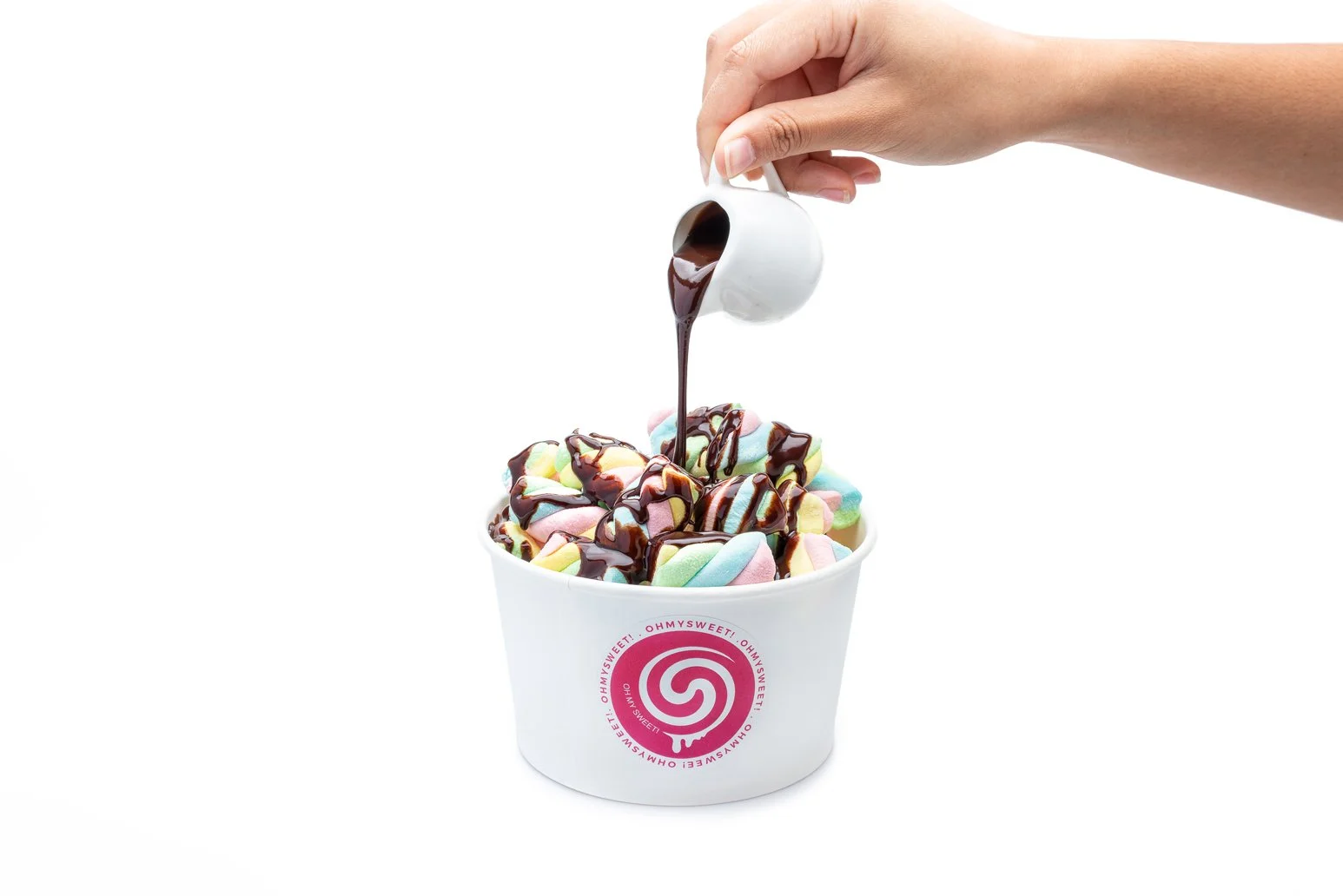 A white cup filled with colorful marshmallows topped with chocolate syrup being poured from a small white pitcher.