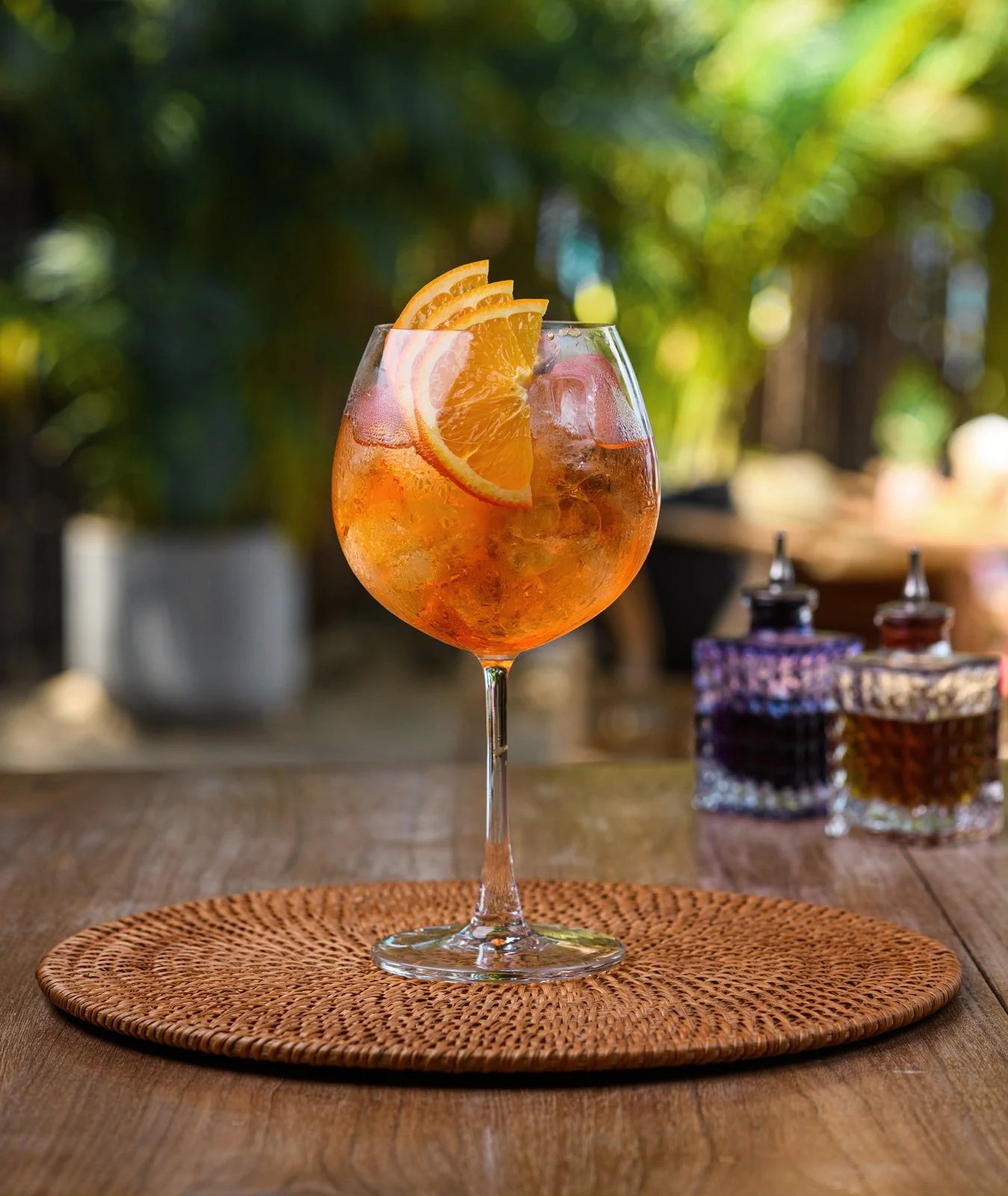 A wine glass filled with an orange-colored drink, garnished with orange slices, sits on a woven placemat on a wooden table. In the background, blurred jars and a tray are visible, with outdoor greenery and sunlight.