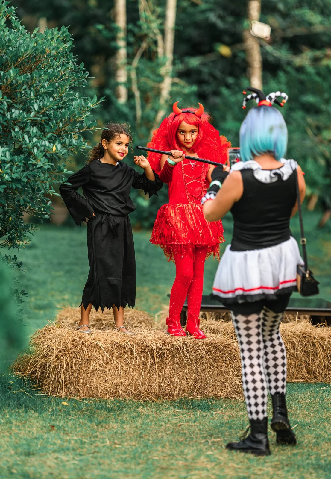 Three children dressed in Halloween costumes outdoors, with two girls standing on a hay bale and a third girl facing away. The girl on the left is dressed as a black cat, the girl in the middle as a devil with red hair, horns, and a red dress, and th