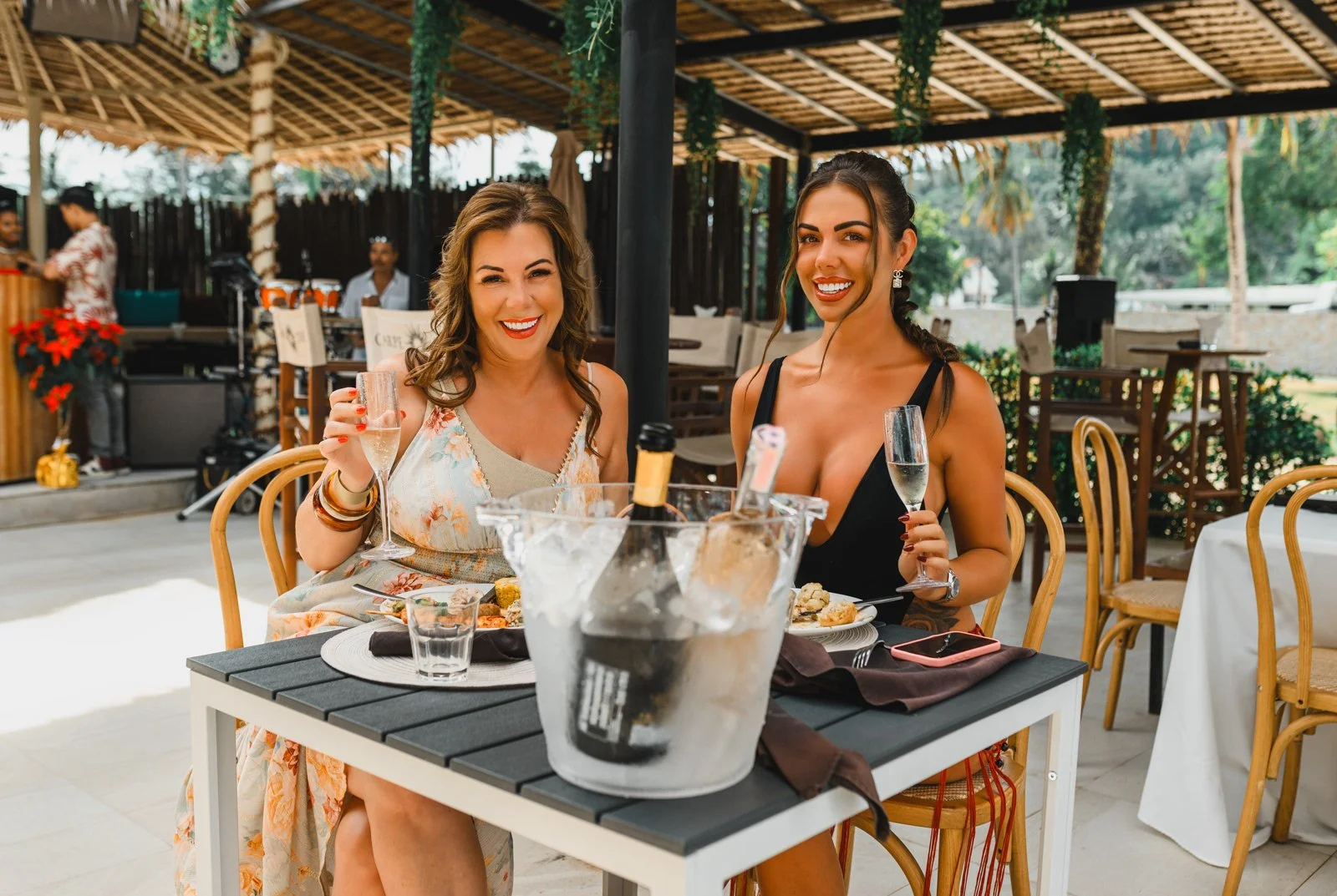 Two women smiling and celebrating at an outdoor restaurant, holding glasses of champagne, with a bucket of ice and wine in front of them.