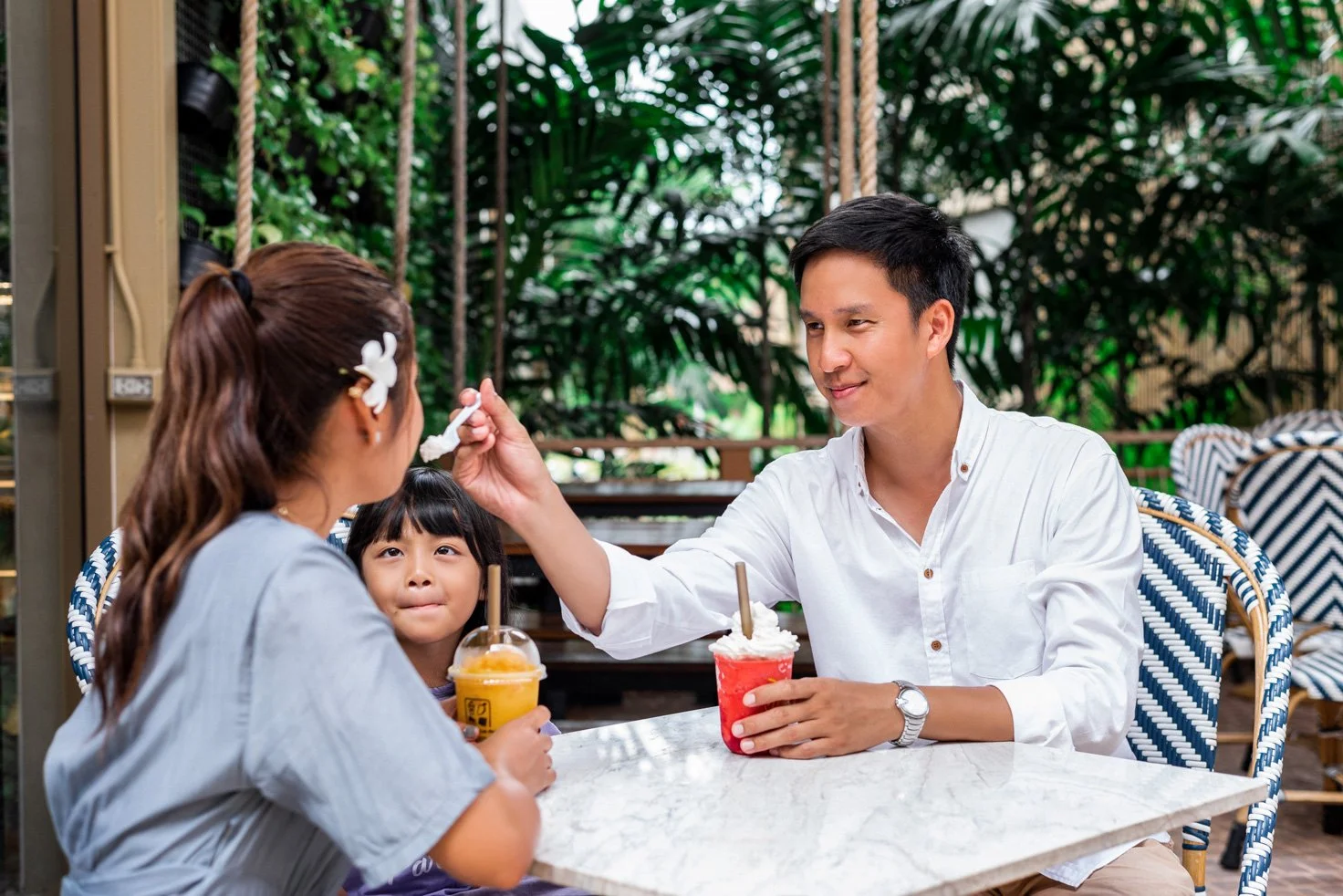 Family sitting at a table in a tropical-themed cafe, with a man feeding a woman a spoonful of dessert, a child watching, and tropical plants in the background.