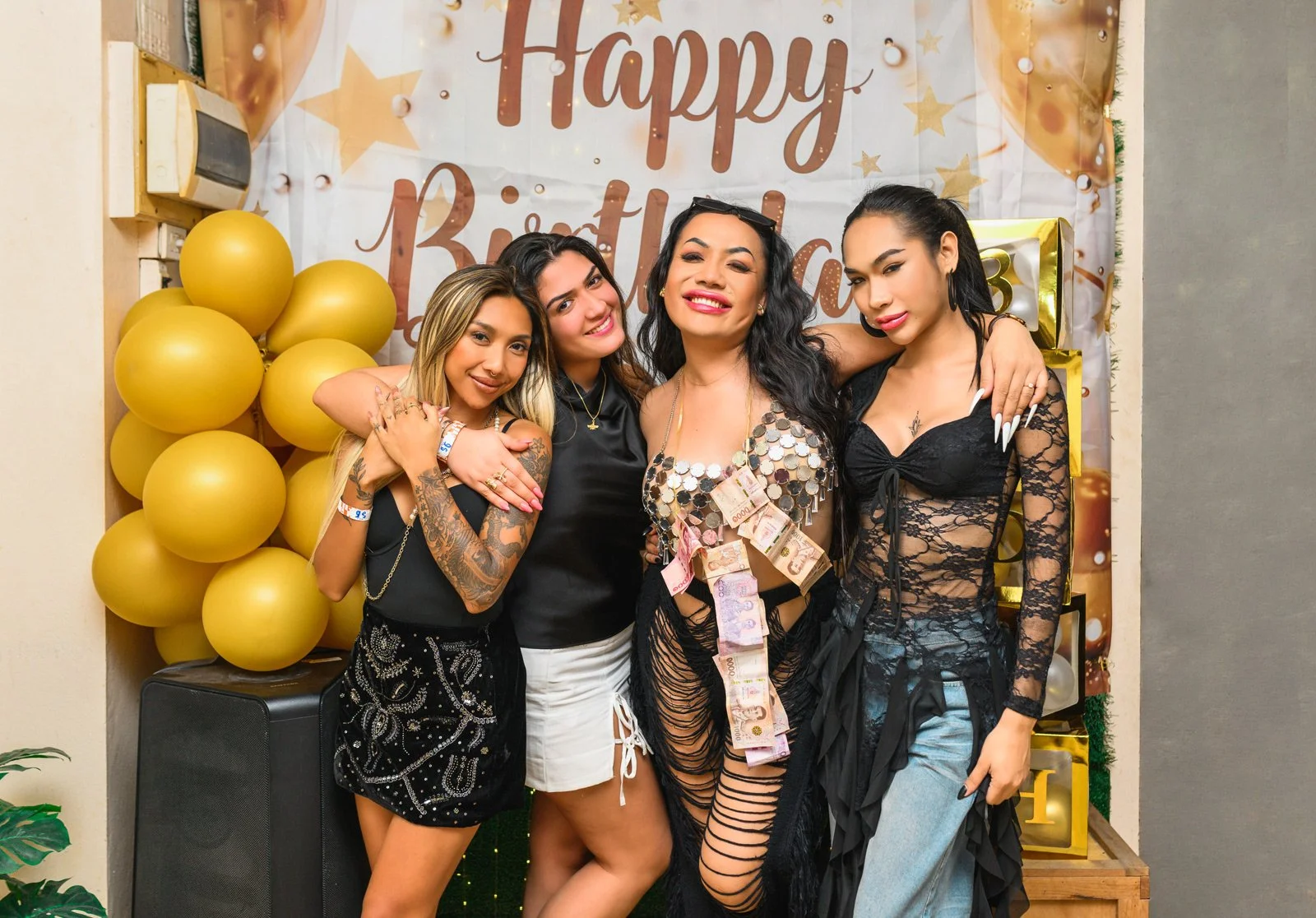 Four women celebrating a birthday in front of a decorated backdrop with balloons and a sign that says "Happy Birthday." The woman in the center is wearing a sequined dress with money pinned on it, and the women are smiling and posing for the photo.