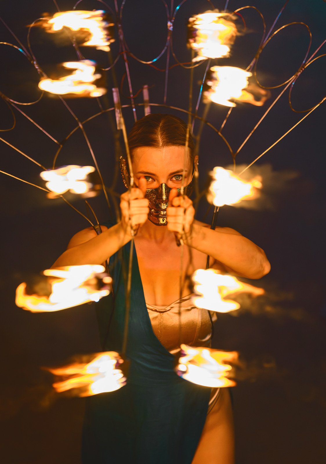 A woman at night holding a sparking firework with flames and sparks surrounding her, wearing a gold mask that covers her nose and mouth.