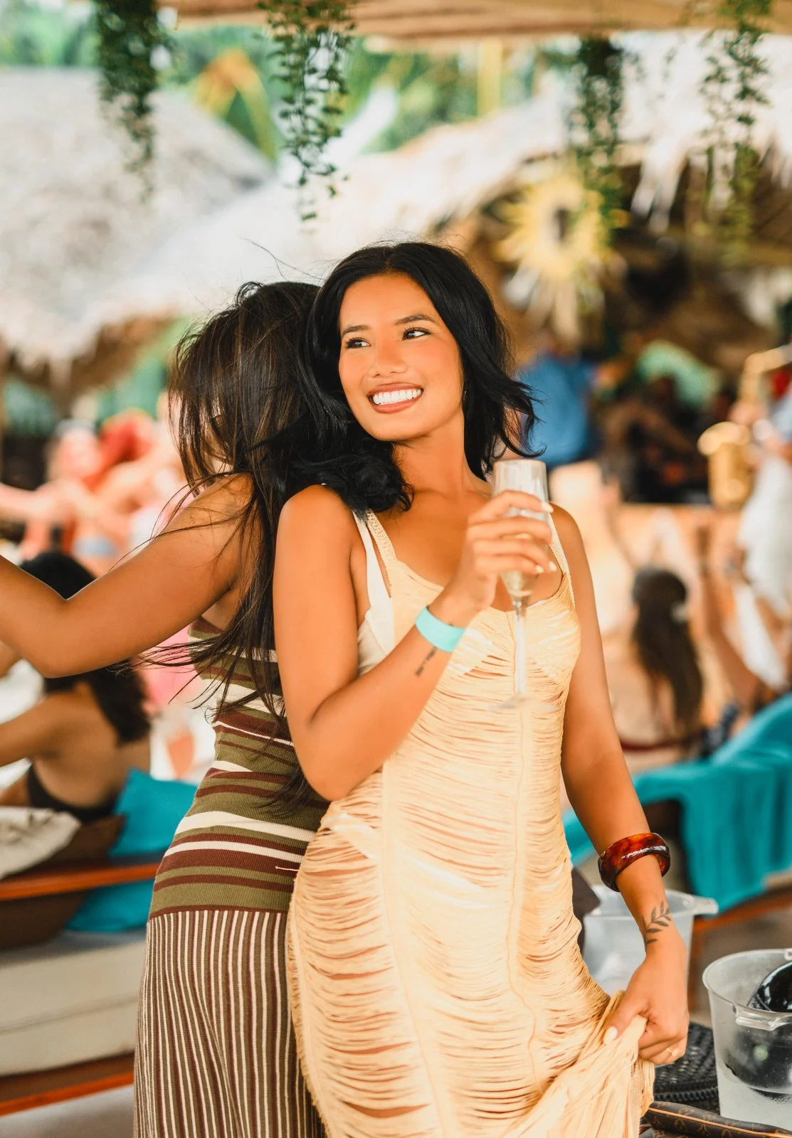 Two women smiling, one holding a glass of champagne at a festive event with greenery and people in the background.