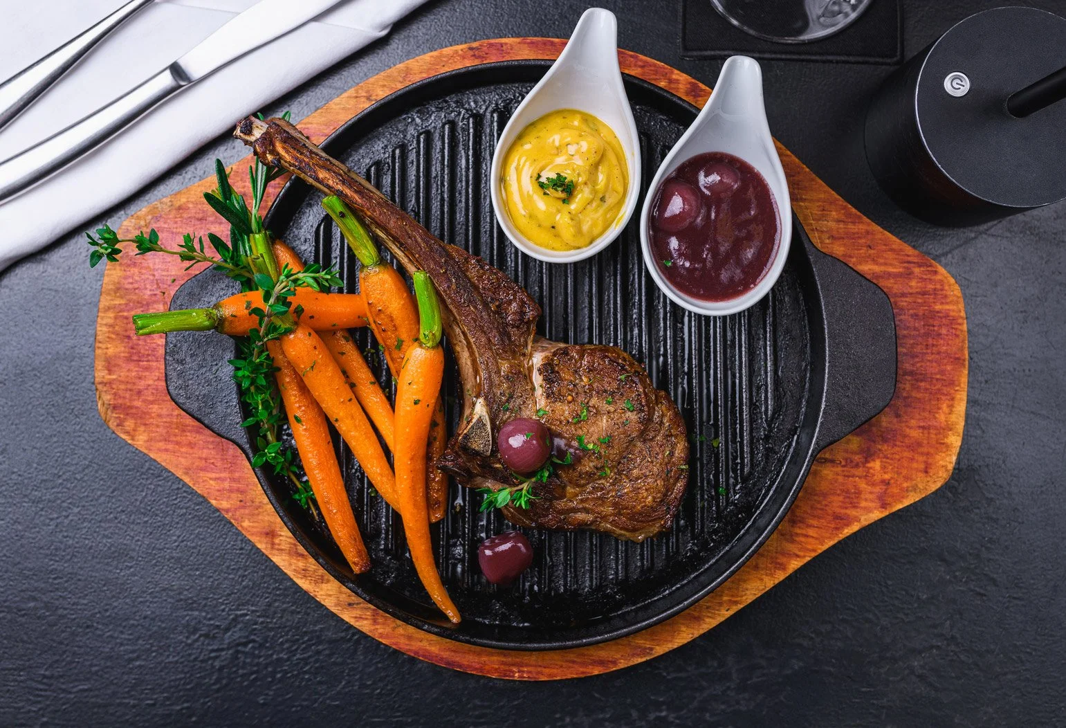 A cast iron skillet with grilled lamb chop, glazed carrots with herbs, and two small bowls of dipping sauces, on a wooden serving board.