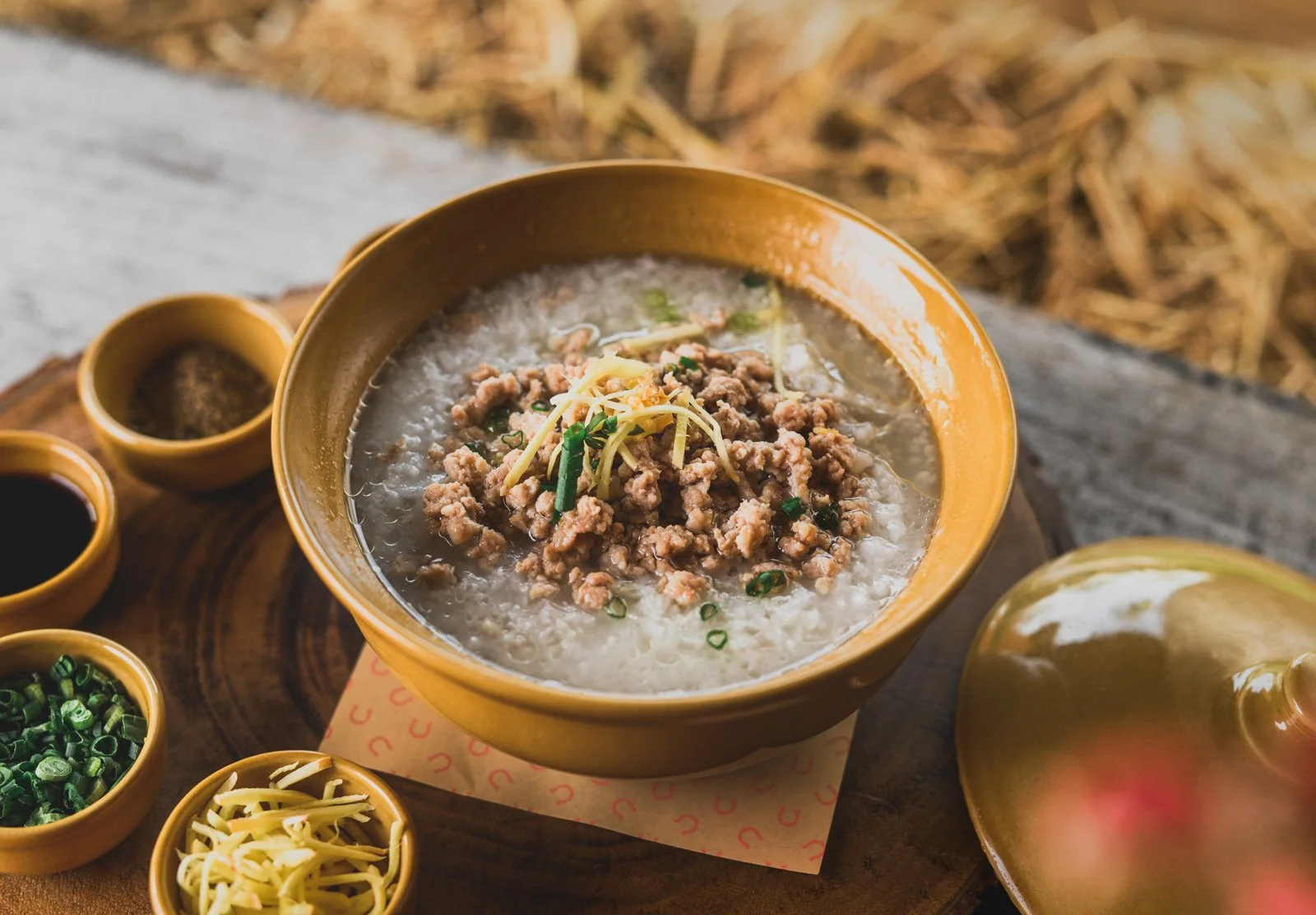A bowl of congee topped with ground meat, ginger, and green onions, surrounded by small bowls of condiments including soy sauce, chopped green onions, shredded ginger, black pepper, and chili paste, on a rustic wooden surface outdoors.